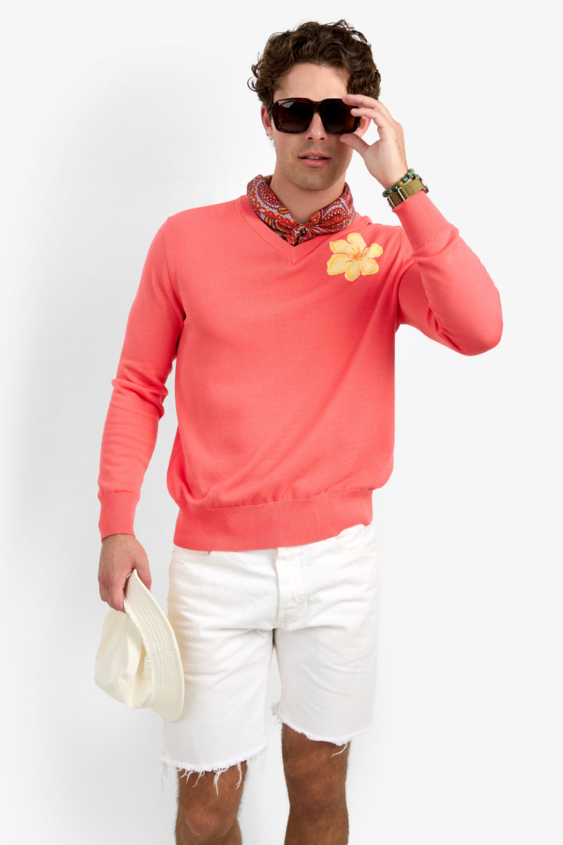 A man wearing the Divino V-Neck Sweater in coral, with a yellow flower, white shorts, sunglasses, a patterned neck scarf, and holding a white hat stands against a plain light background.