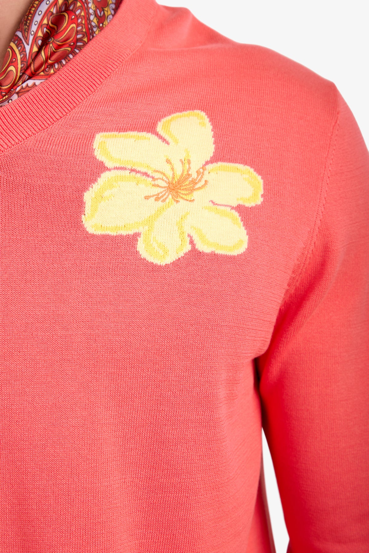 A close-up of the Divino V-Neck Sweater in coral pink, featuring a bold yellow flower on the chest, styled with a patterned red scarf.