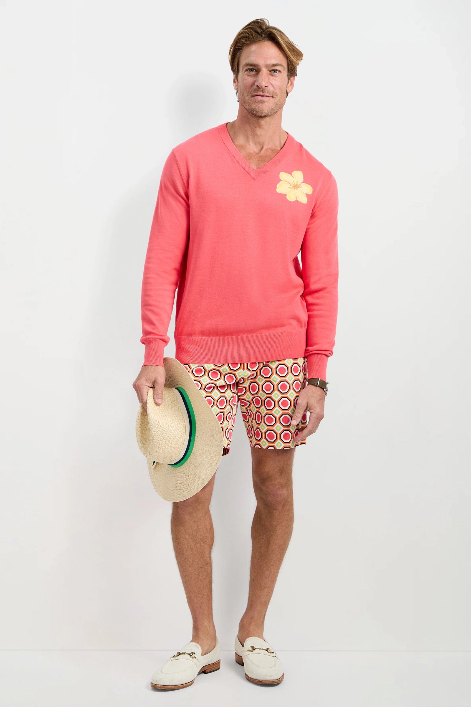 A man stands against a white background wearing the Divino V-Neck Sweater in Corso Coral with a yellow flower detail, patterned shorts, white loafers, and holding a straw hat in his left hand.