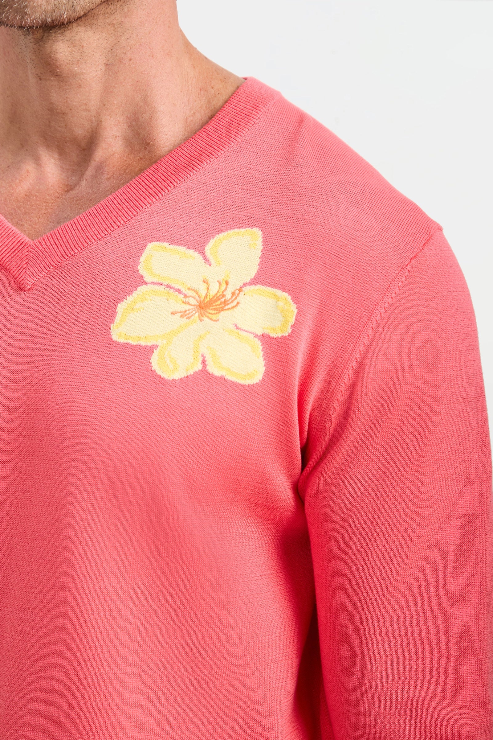 A close-up of someone wearing the Divino V-Neck Sweater in pink, which features a large yellow flower design on the left shoulder. Only the upper chest, shoulder, and neck are shown.
