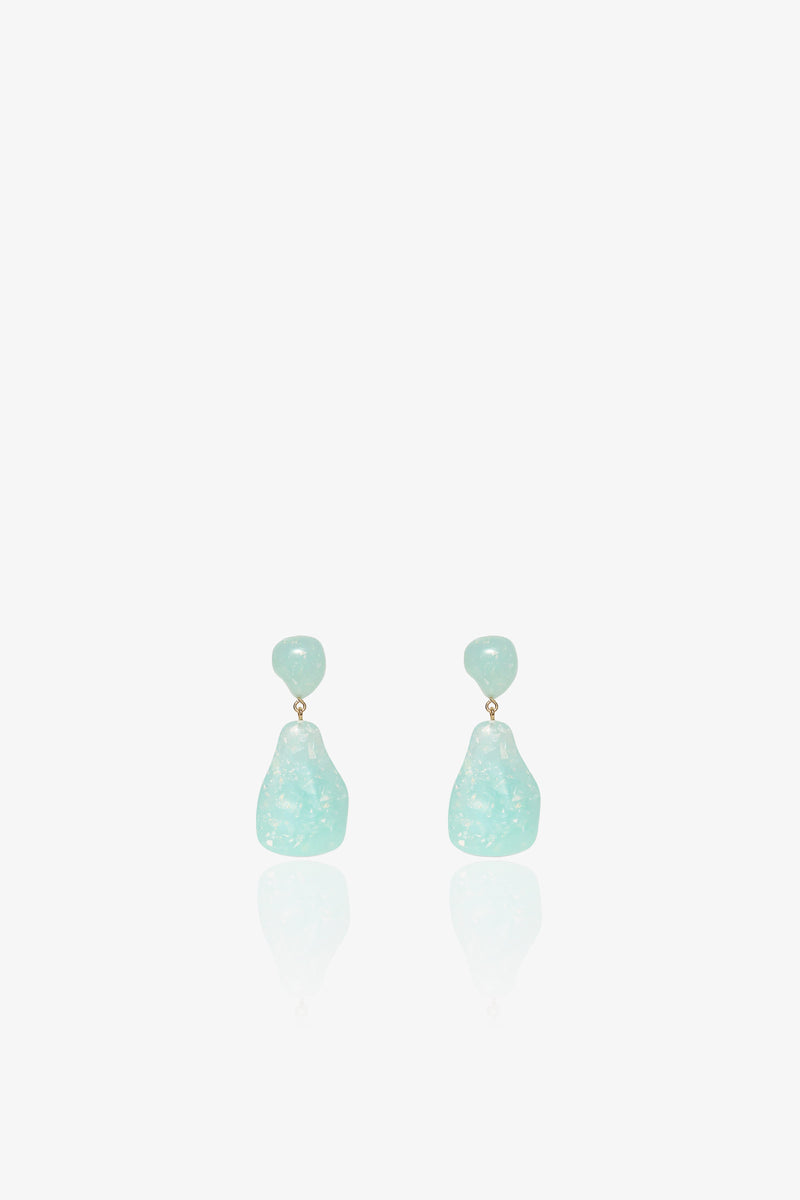 The Wilma Earrings feature small, round, light blue studs and larger, irregularly shaped translucent blue drops in a dangling design, displayed against a white background.