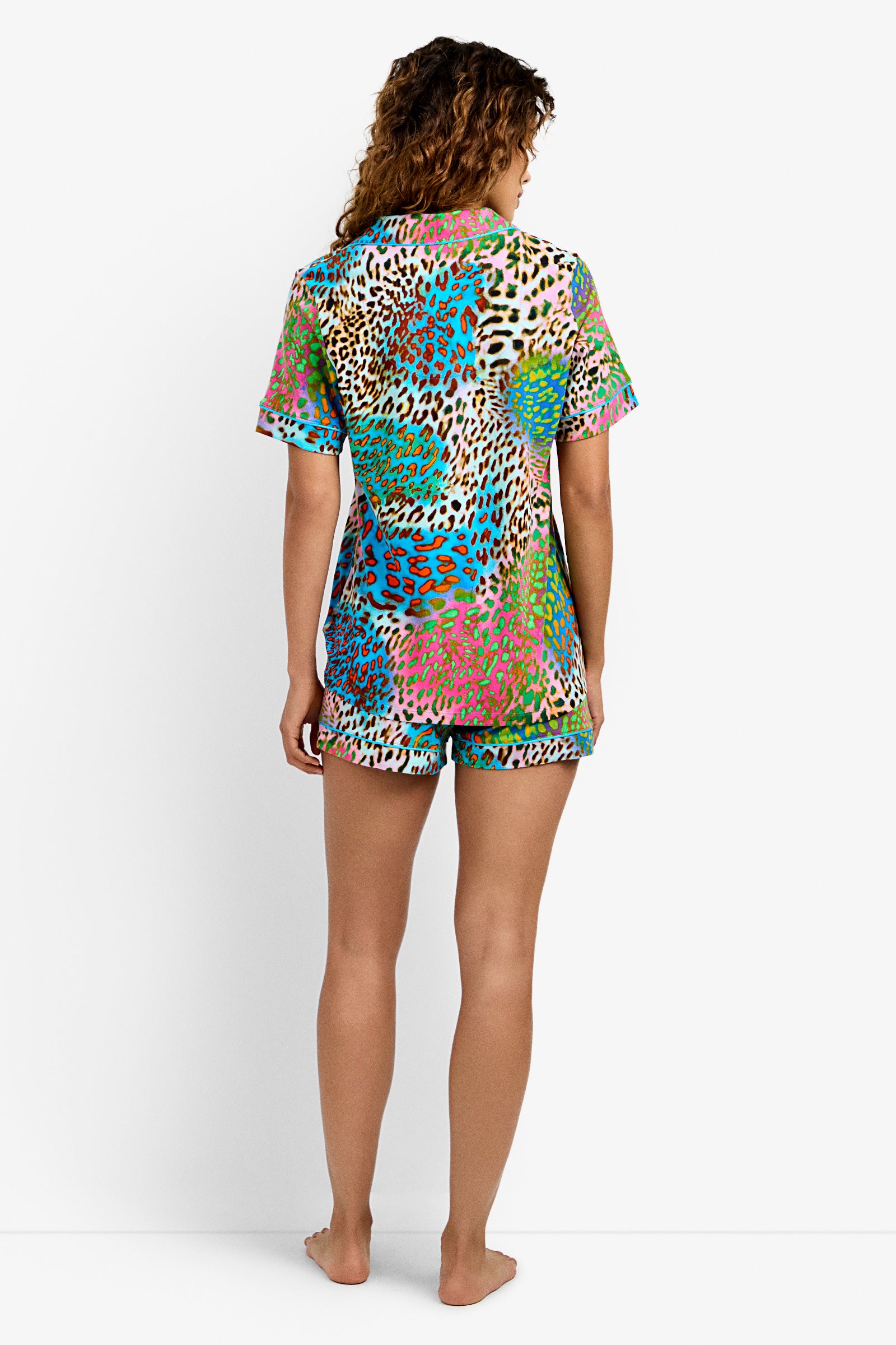 A woman stands barefoot facing away, wearing the Shorty PJ Set - Spray Leopard, a colorful short-sleeve pajama set featuring a vibrant multicolored leopard print pattern. The background is plain white.