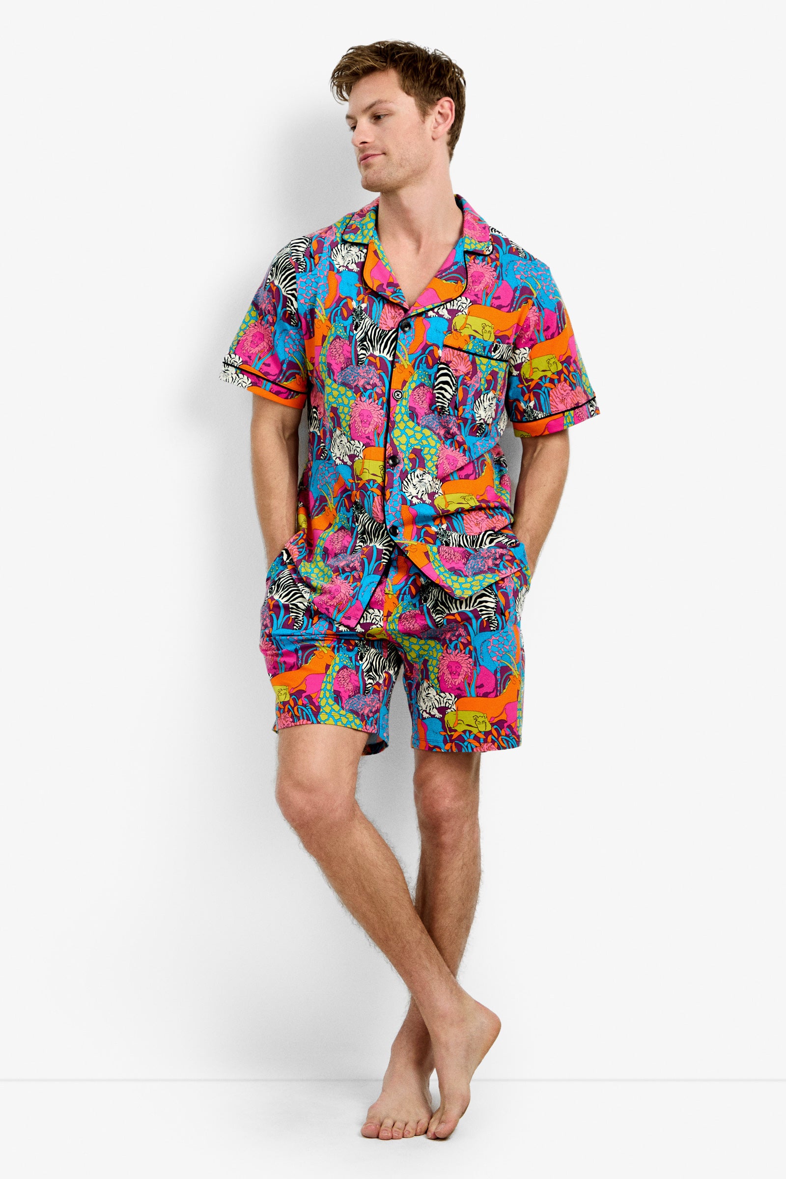 A man stands barefoot against a white background, wearing the Men's Boxer PJ Set - Jungle, which features a matching colorful pajama set with a bold, tropical animal print in bright pink, blue, orange, and purple. His hands are in his shorts’ pockets.