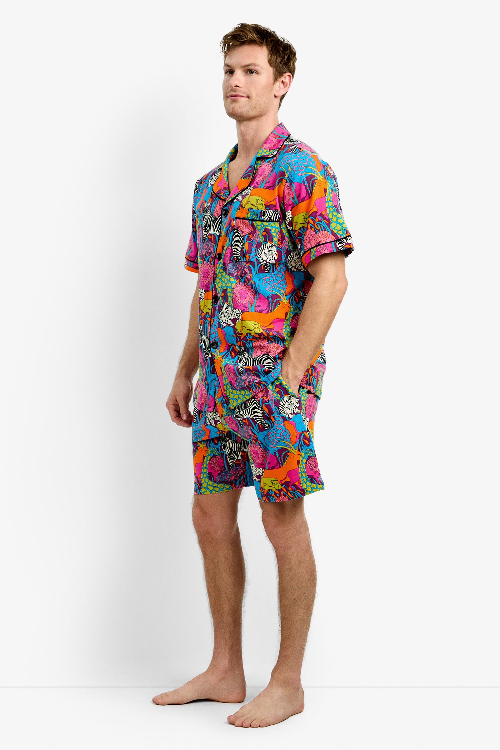 A man stands barefoot against a plain white background, wearing the Men's Boxer PJ Set - Jungle, a colorful matching short-sleeve shirt and shorts featuring a vibrant jungle-inspired print.