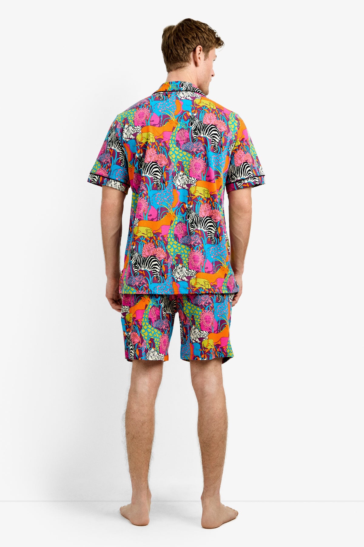 A man stands barefoot facing away, wearing the Men's Boxer PJ Set - Jungle, featuring a vibrant animal print with zebras and other wildlife on a blue background.