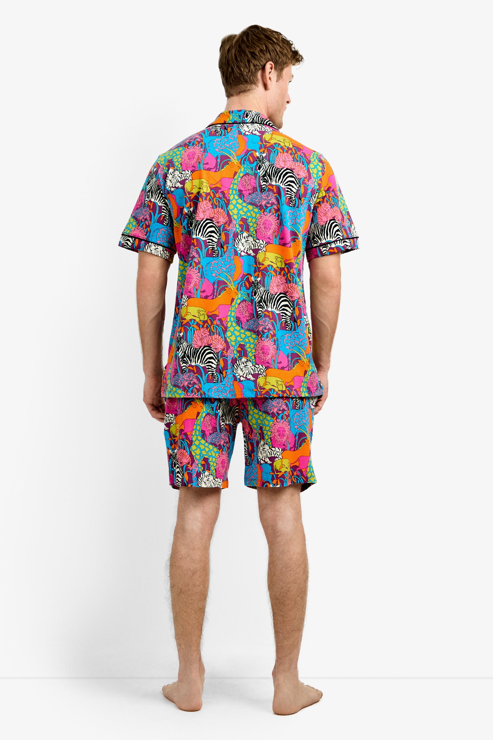 A man stands barefoot facing away, wearing the Men's Boxer PJ Set - Jungle, featuring a vibrant animal print with zebras and other wildlife on a blue background.