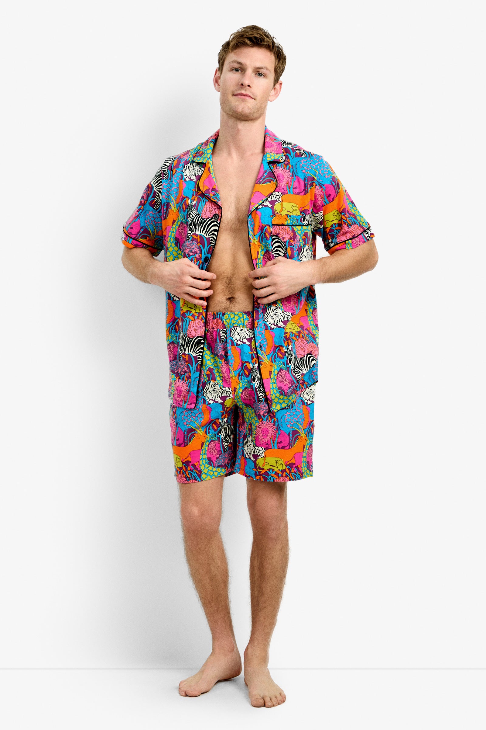 A man stands barefoot against a white background, wearing the Men's Boxer PJ Set - Jungle, which features a colorful animal print in pink, blue, orange, and yellow. The set includes a short-sleeve button-up shirt and matching shorts.