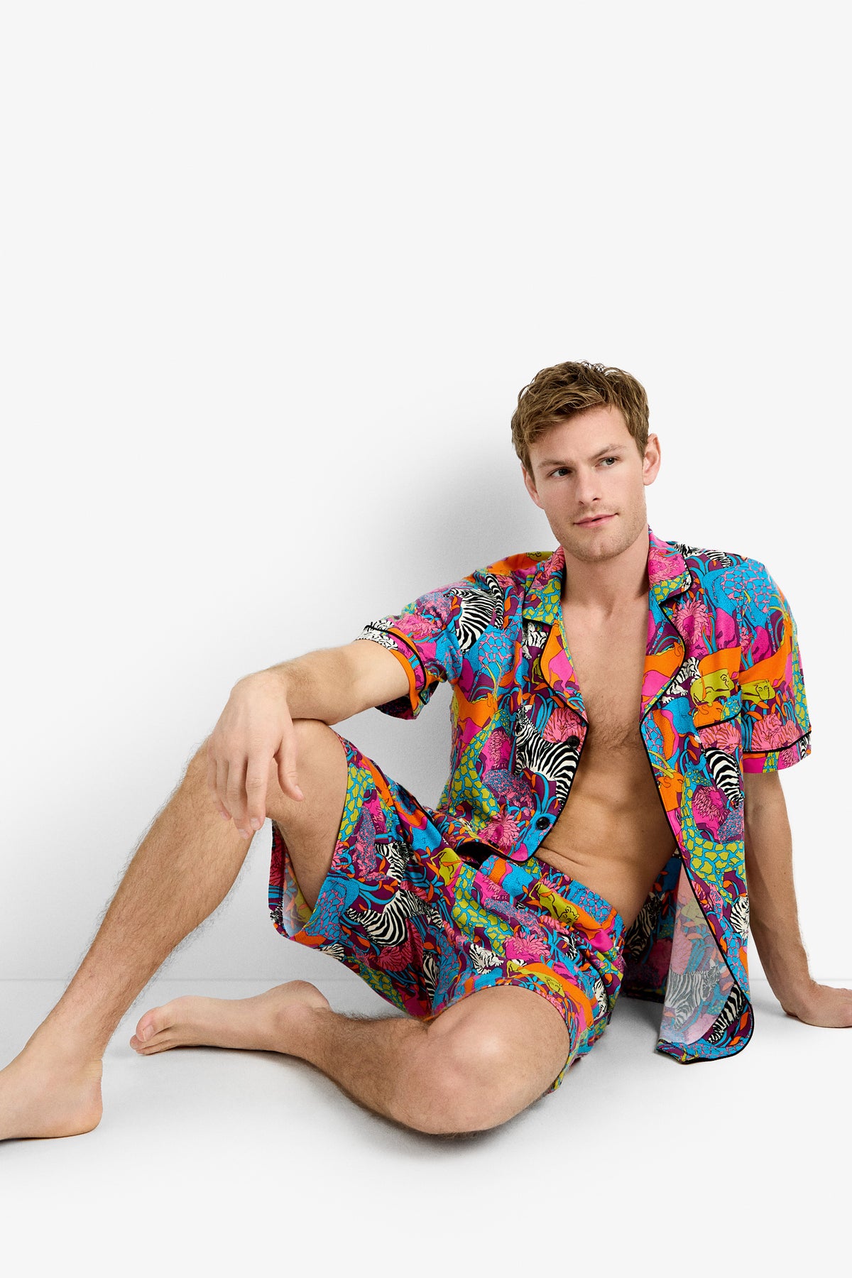 A man sits on the floor against a white background, wearing the Men's Boxer PJ Set - Jungle, featuring a colorful matching short-sleeve shirt and shorts with vibrant jungle-inspired patterns.