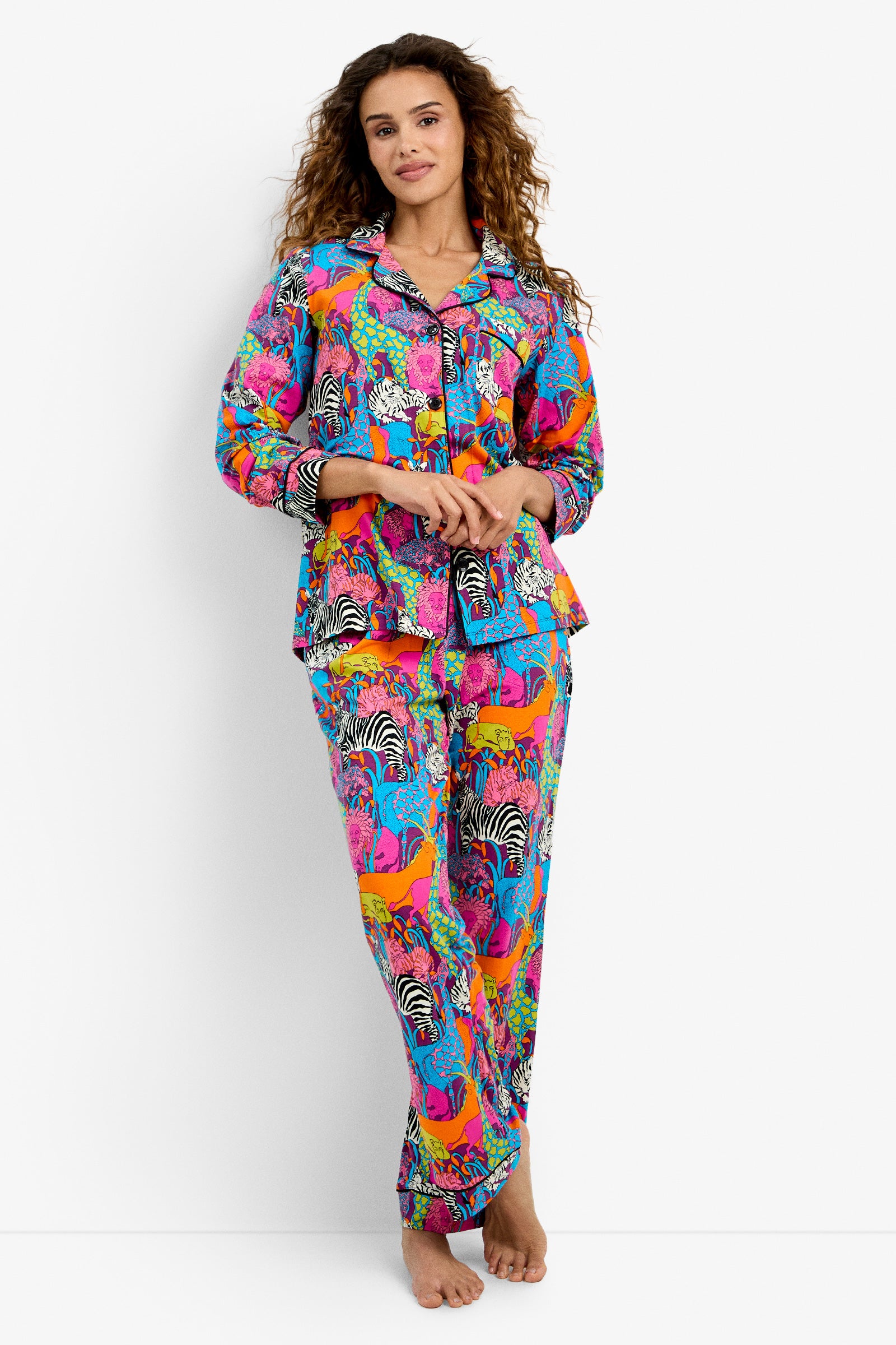 A woman with long, curly hair stands barefoot against a white background, wearing the Classic PJ Set - Jungle, featuring colorful animal patterns in bright pink, blue, orange, and yellow. She looks relaxed and comfortable.