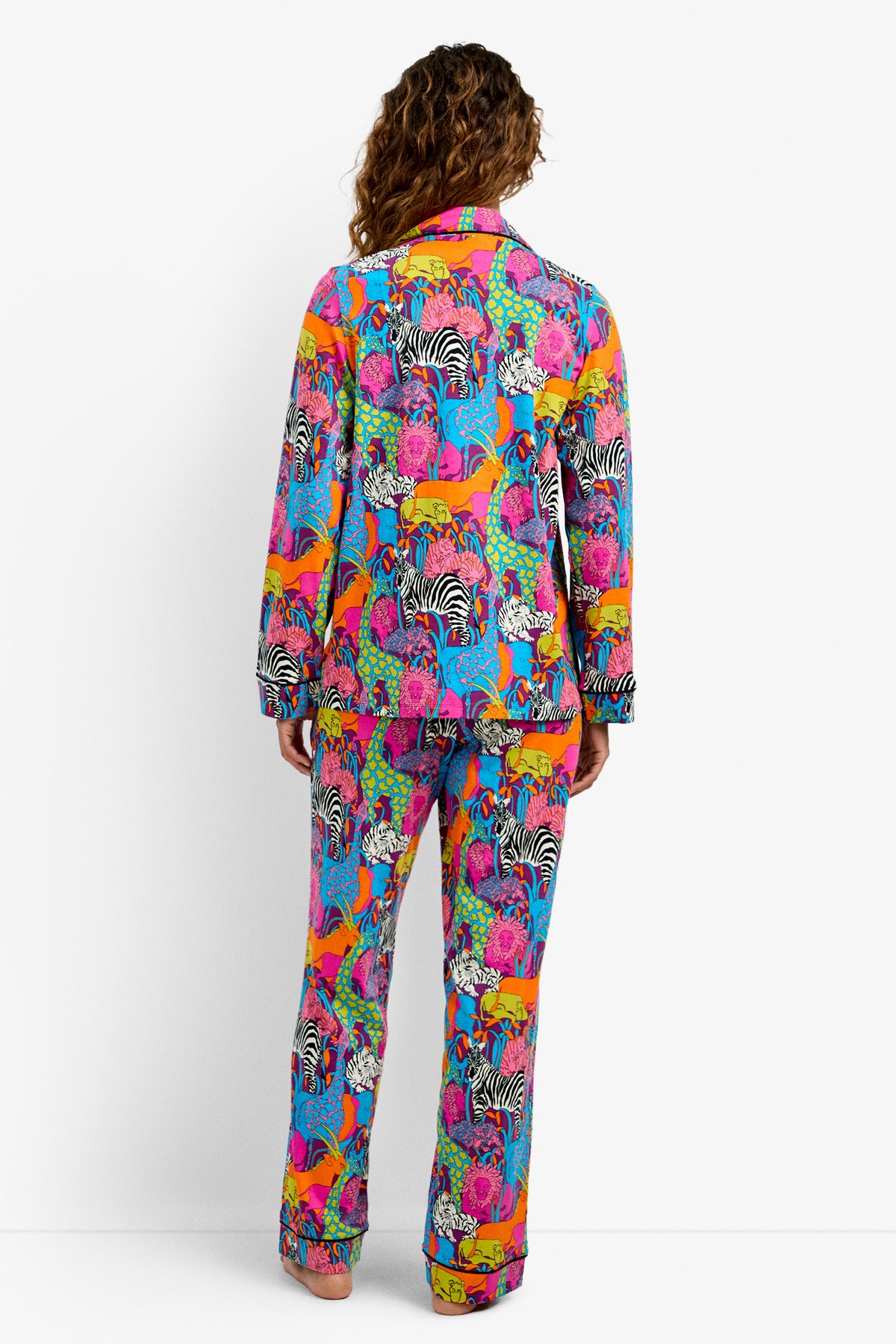 Someone stands facing away, wearing the Classic PJ Set - Jungle, featuring a vibrant animal pattern with zebras, tigers, and more in pink, orange, blue, green, and yellow on a white background.