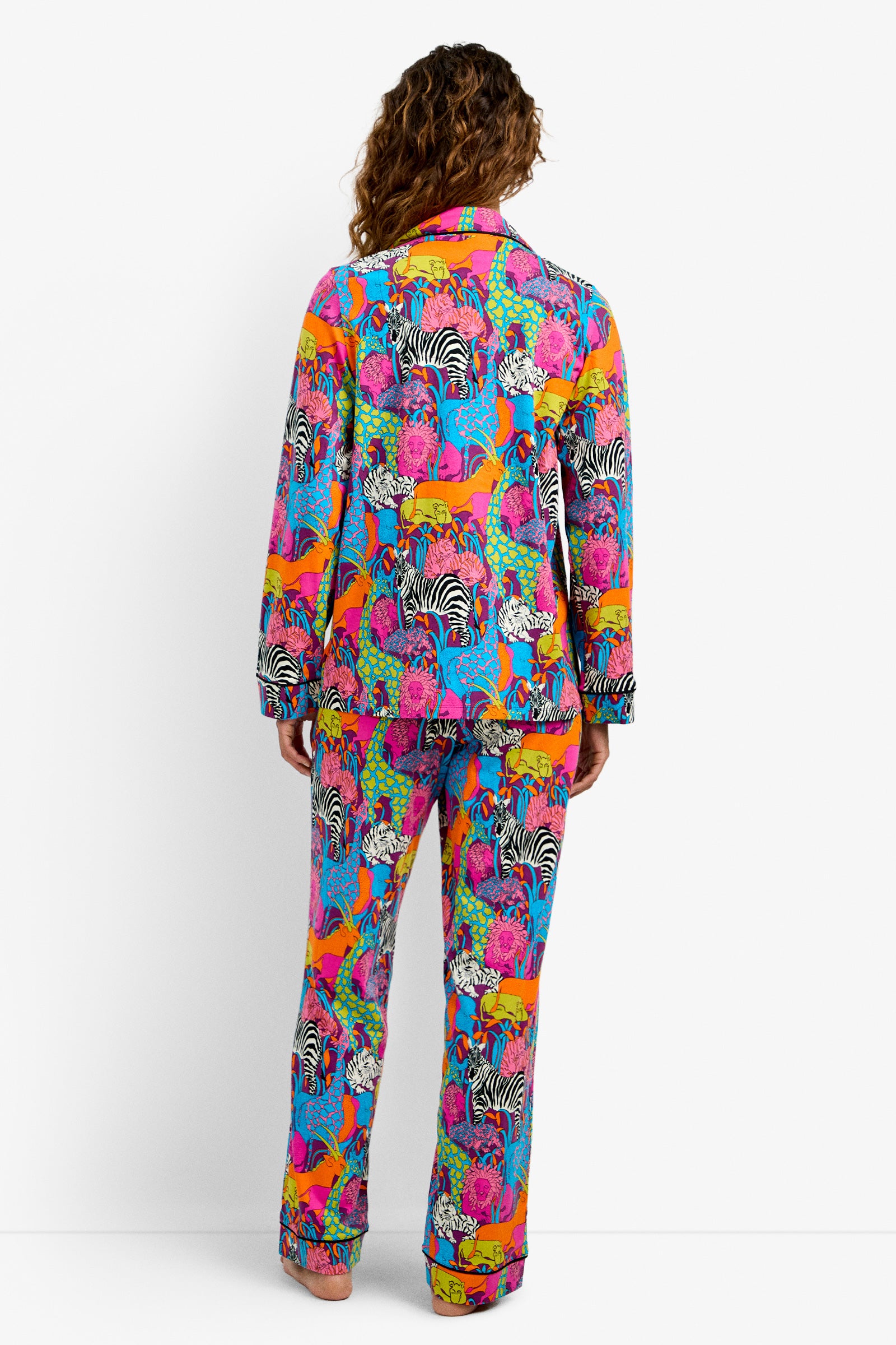 Someone stands facing away, wearing the Classic PJ Set - Jungle, featuring a vibrant animal pattern with zebras, tigers, and more in pink, orange, blue, green, and yellow on a white background.