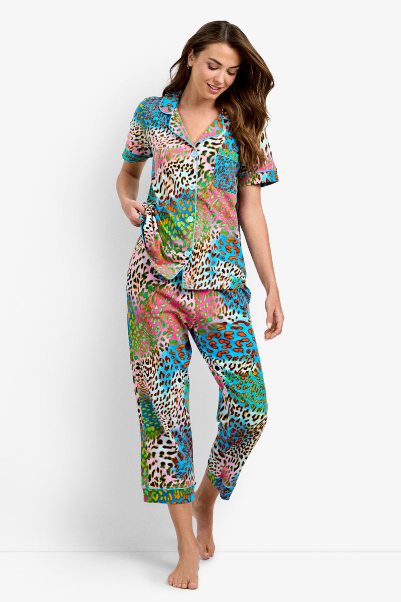 A woman smiles wearing the Crop Classic PJ Set - Spray Leopard, featuring a short-sleeve button-up top and matching pants in colorful blue, green, pink, and brown leopard print. She walks barefoot against a white background.