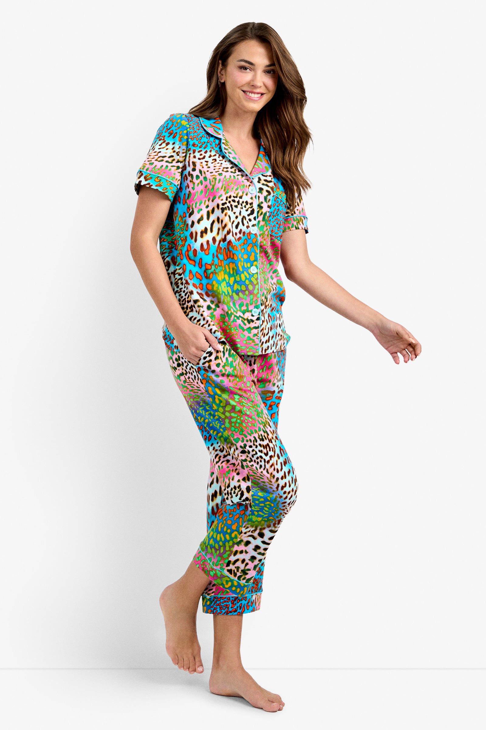 A smiling woman with long brown hair wears the Crop Classic PJ Set in Spray Leopard, featuring a colorful leopard print, short sleeves, and cropped pants. She stands barefoot against a plain white background.