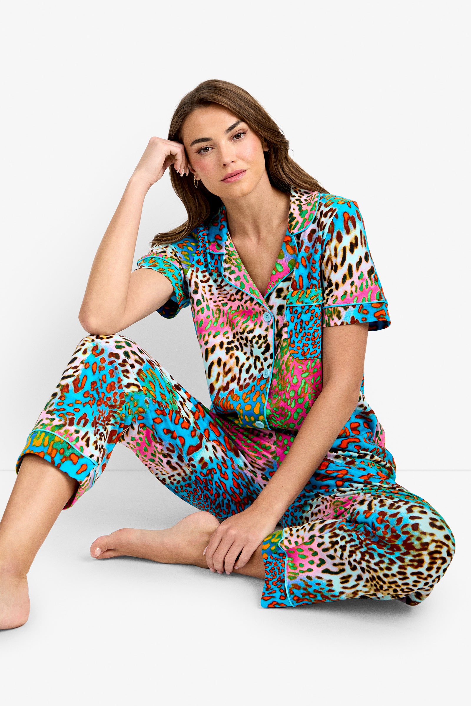 A woman with long brown hair sits barefoot on the floor, wearing the Crop Classic PJ Set in Spray Leopard—a colorful leopard-print pajama set—against a white background, resting her head on one hand and looking at the camera.