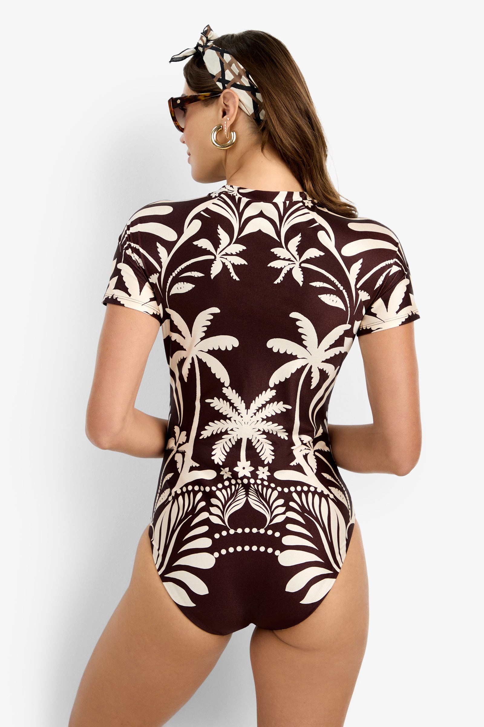 A woman faces away wearing the Short-Sleeve Zip-Up Paddlesuit, featuring a brown and white palm tree pattern. She accessorizes with sunglasses, a headscarf, and large hoop earrings.