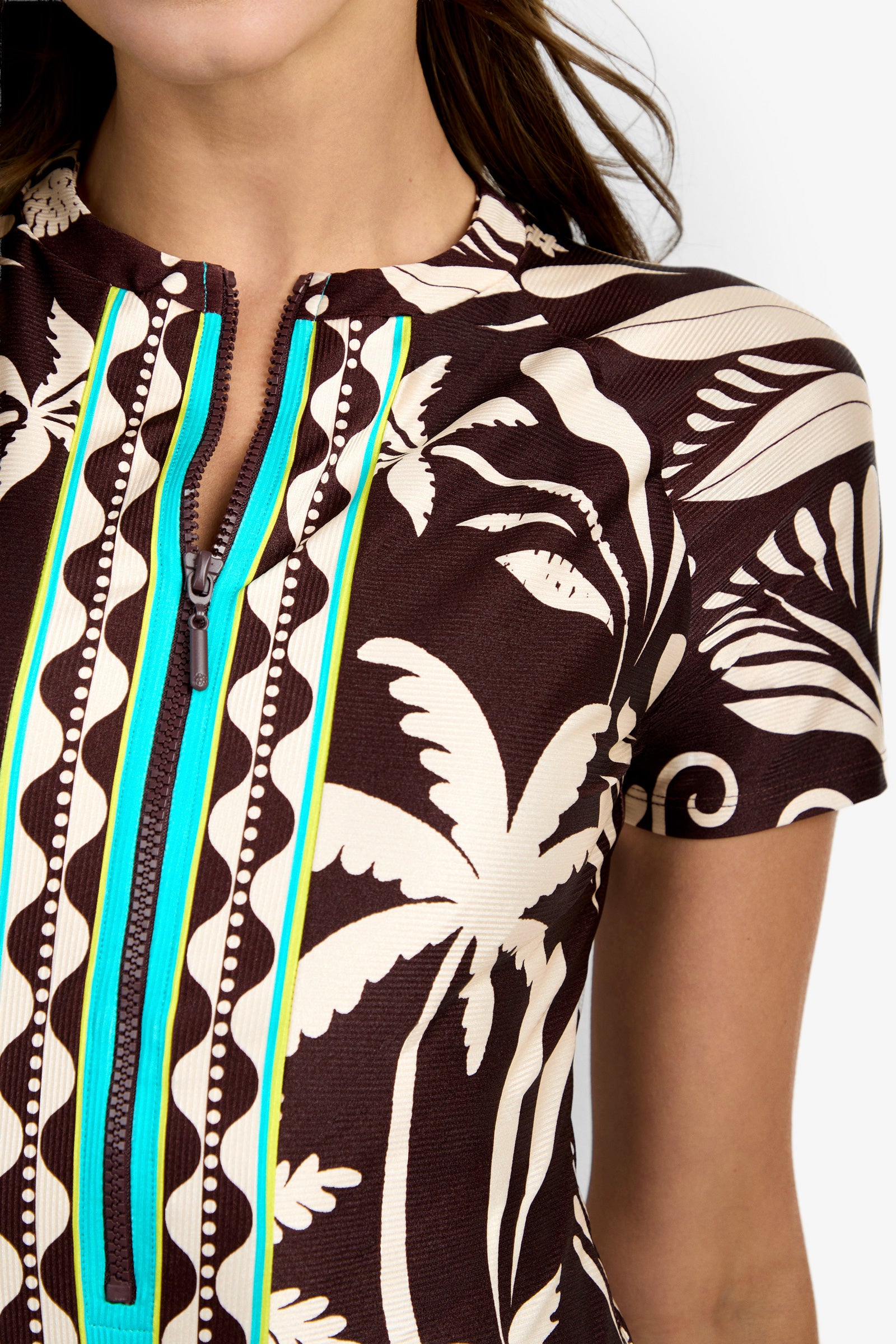 A close-up of a woman wearing the Short-Sleeve Zip-Up Paddlesuit, featuring brown fabric with white tropical leaf and palm prints, blue and yellow trim, and a front zipper. Only part of her face and upper body are shown.