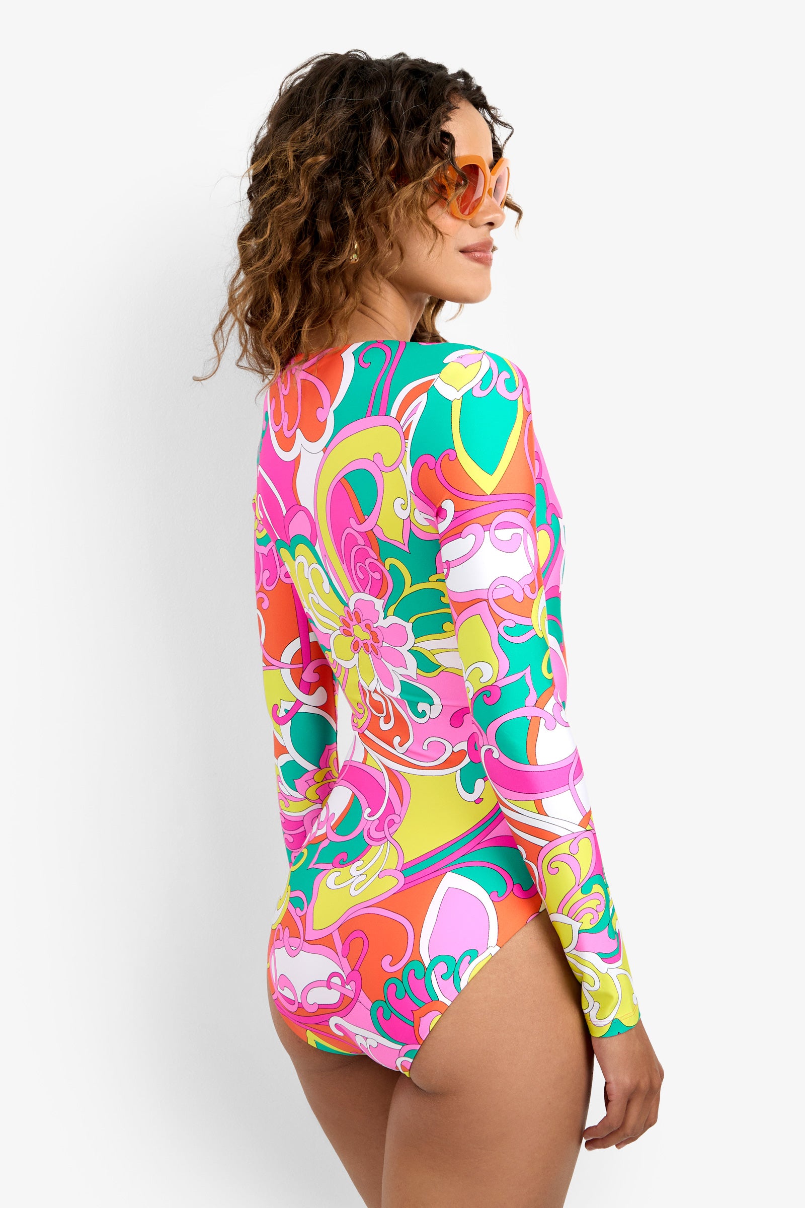 A woman with curly hair and orange sunglasses models the Long-Sleeve Ring Paddlesuit, a colorful floral swimsuit, standing slightly turned from the camera against a plain white background.