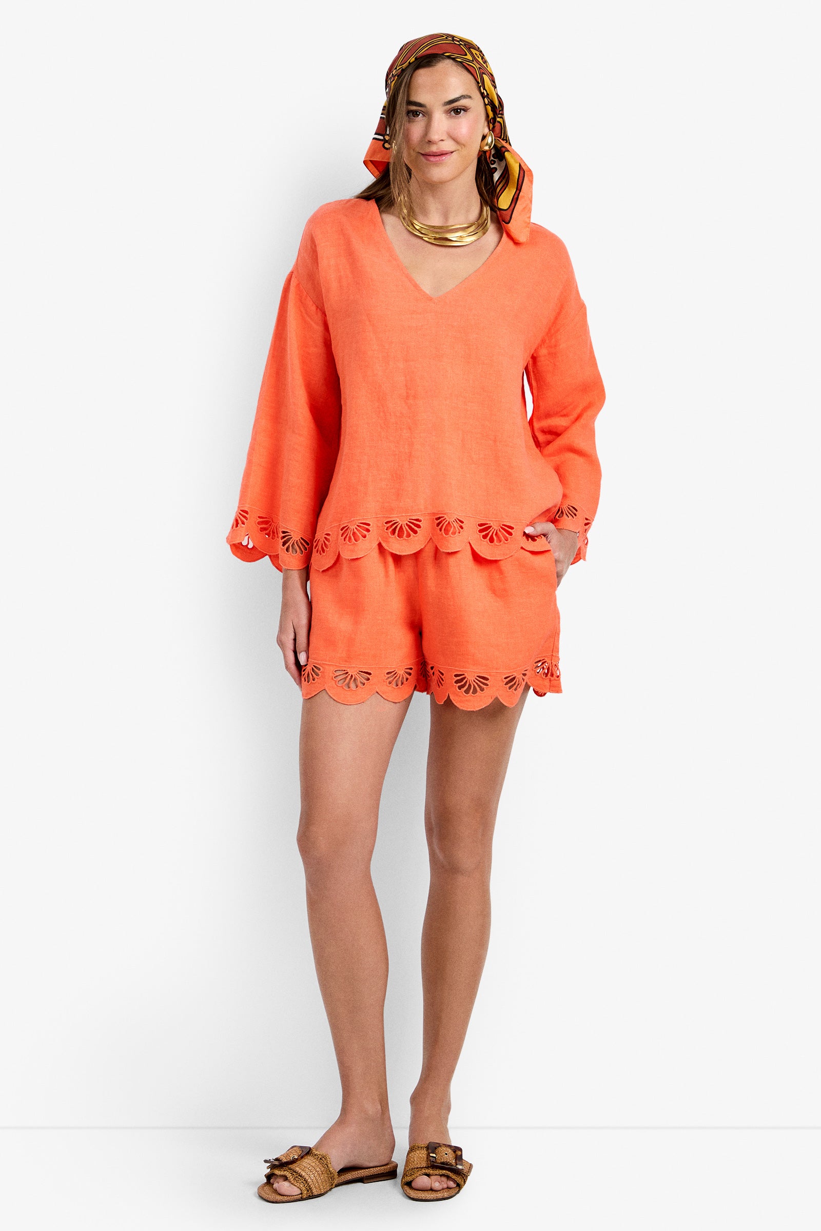 A woman smiles, wearing the Mar Top in CALYPSO CORAL with scalloped edges and matching shorts, paired with a patterned headscarf, chunky necklace, and sandals.