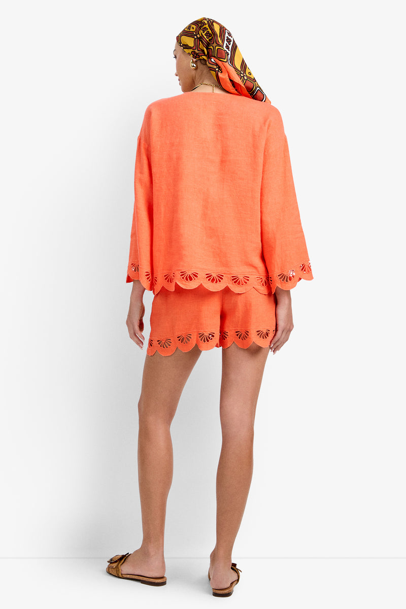 A person stands facing away, wearing the Mar Top—a loose-fitting, coral top paired with matching shorts featuring scalloped cut-out hems. They complete the look with a colorful headscarf and flat sandals.