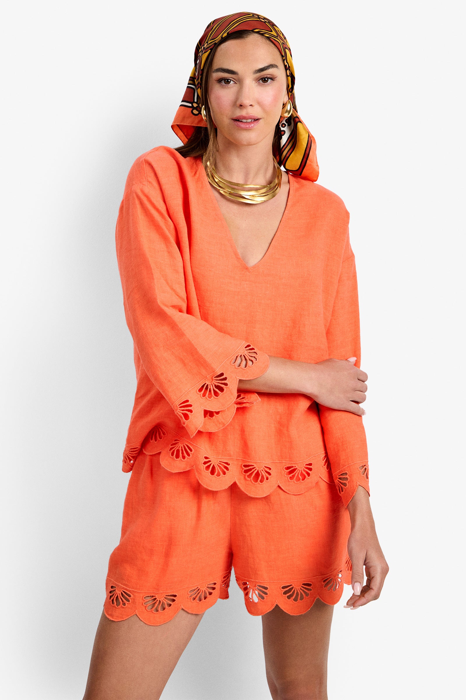A woman wears the Mar Top, an orange linen top with scalloped, cutout edges, paired with matching shorts. She accessorizes with a gold necklace, hoop earrings, and a colorful striped headscarf.