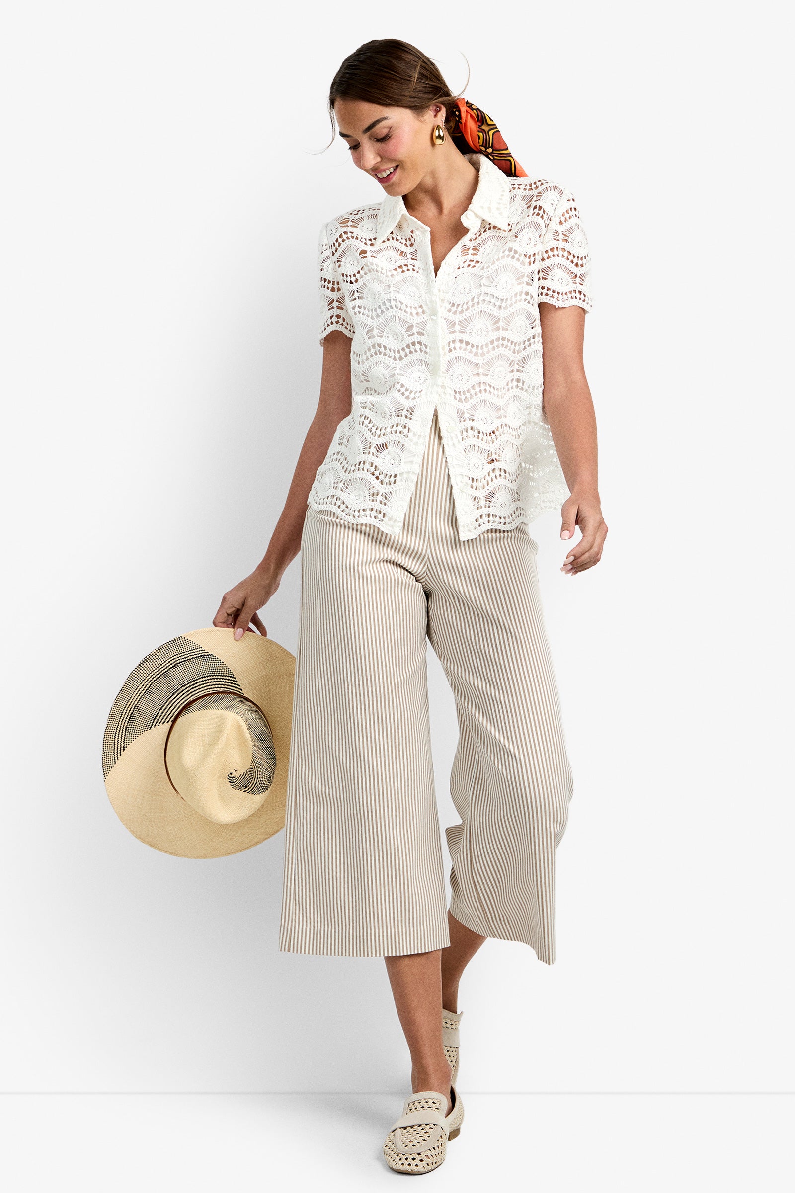 Wearing the Triton Top in WHITEWASH, a woman paired it with beige and white striped wide-leg cropped pants, woven light shoes, and a colorful scarf in her hair as she smiles and looks down, holding a large floppy hat.