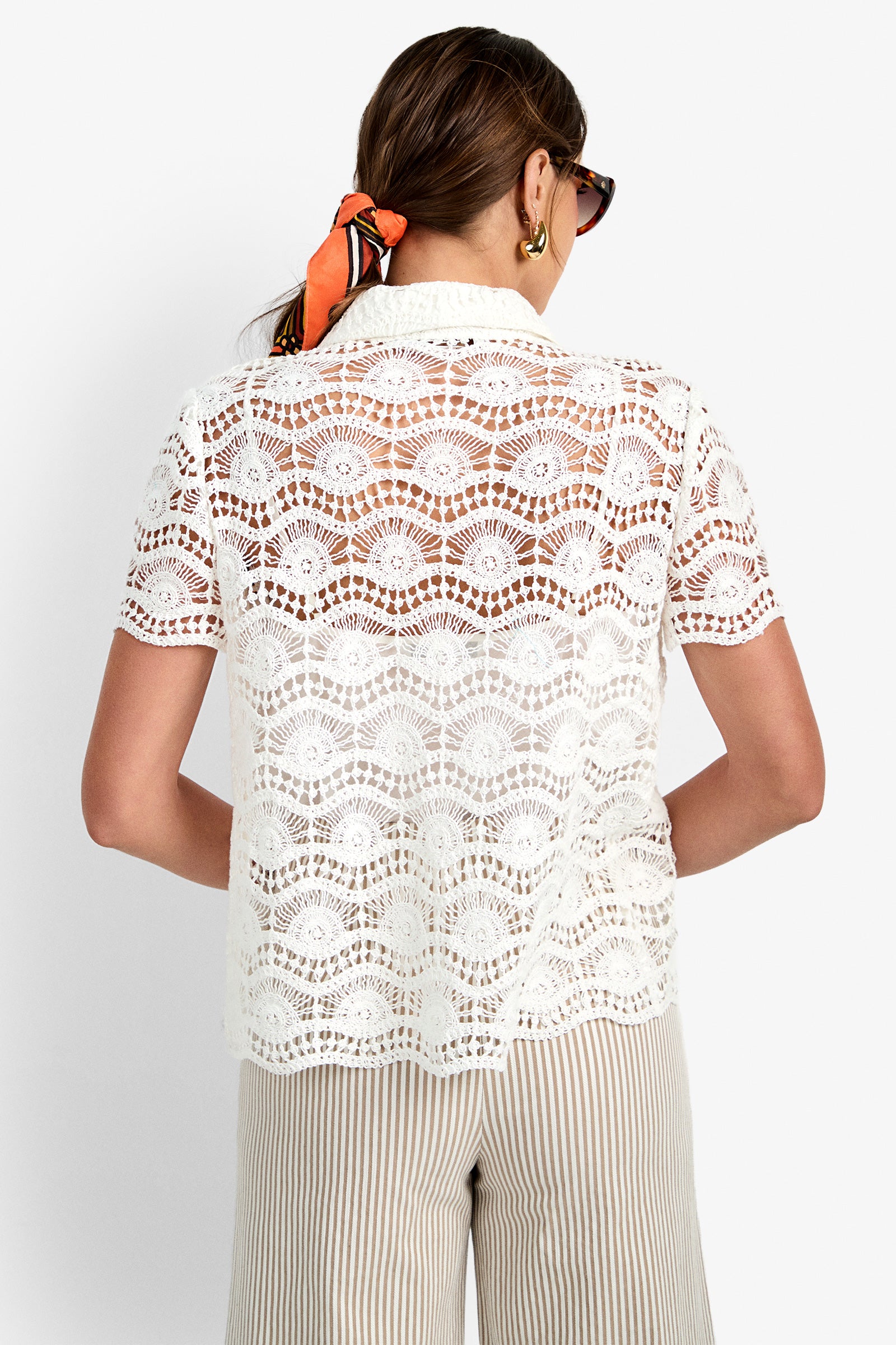 A woman with sunglasses and a hair scarf is shown from the back, wearing the sheer white lace short-sleeve Triton Top and light-colored striped pants.