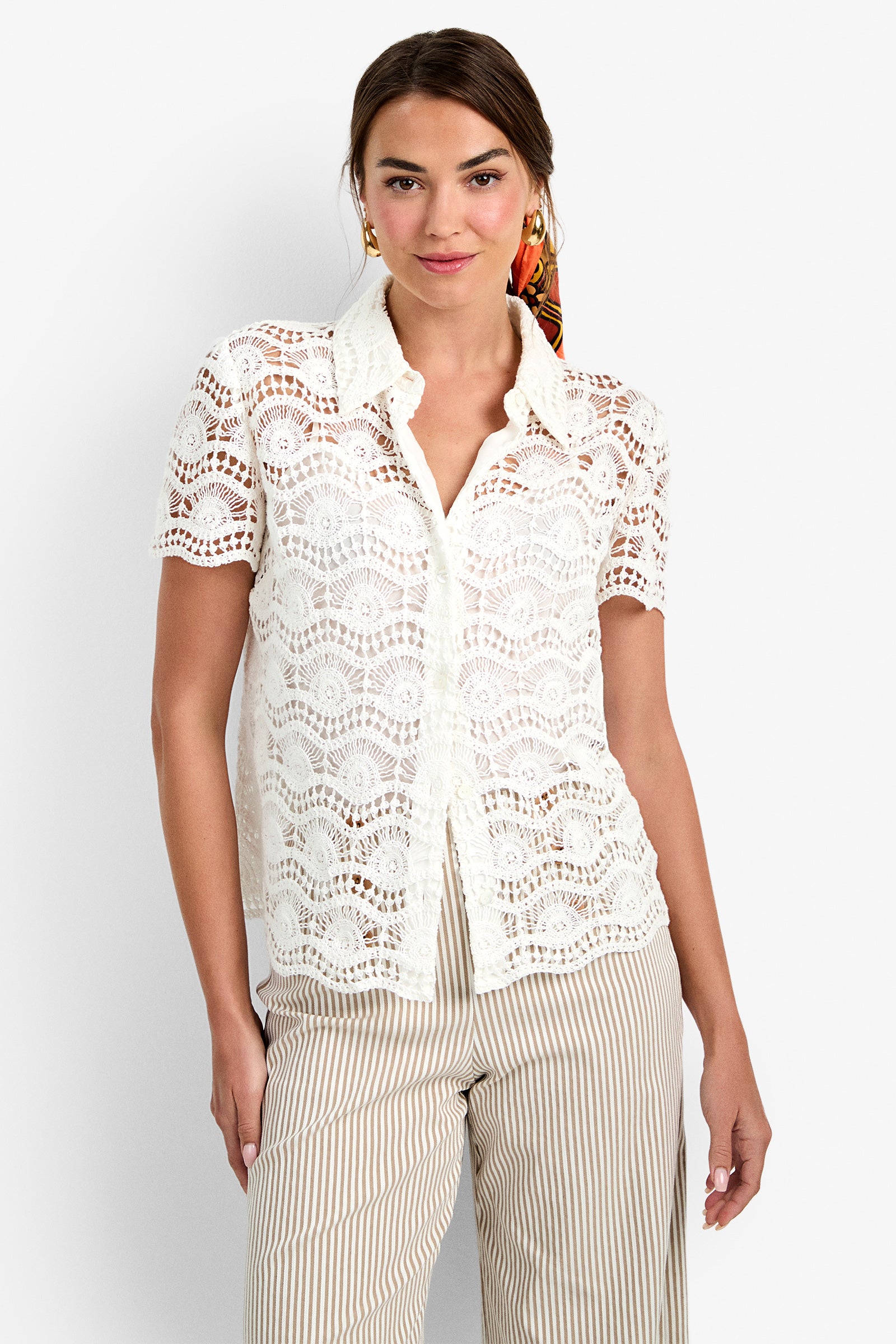 A woman smiles in the Triton Top, a white short-sleeve collared lace blouse with scalloped patterns, paired with beige and white striped pants, against a plain white background.