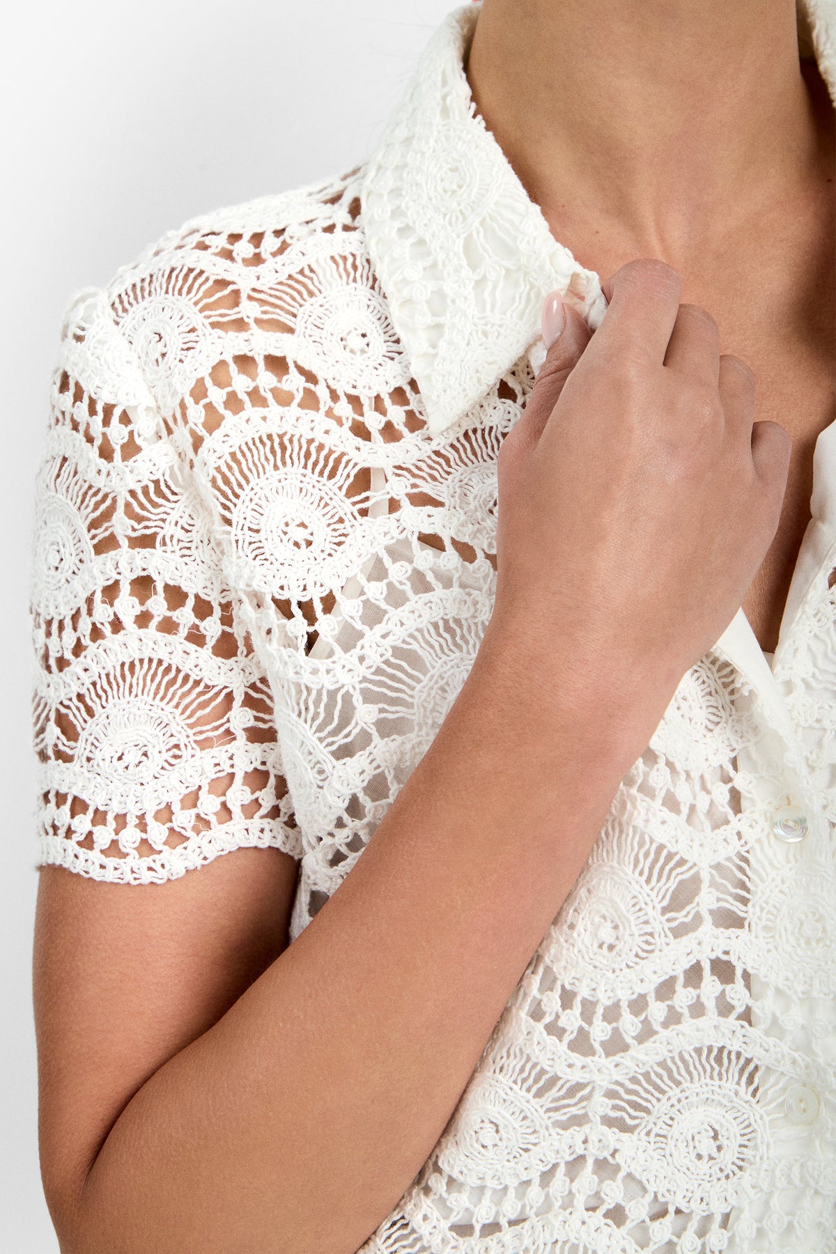 A person wears the Triton Top, a white short-sleeve crochet lace blouse with a collar, showcasing its intricate circular and wave-like patterns while holding the neckline. The background is plain white.