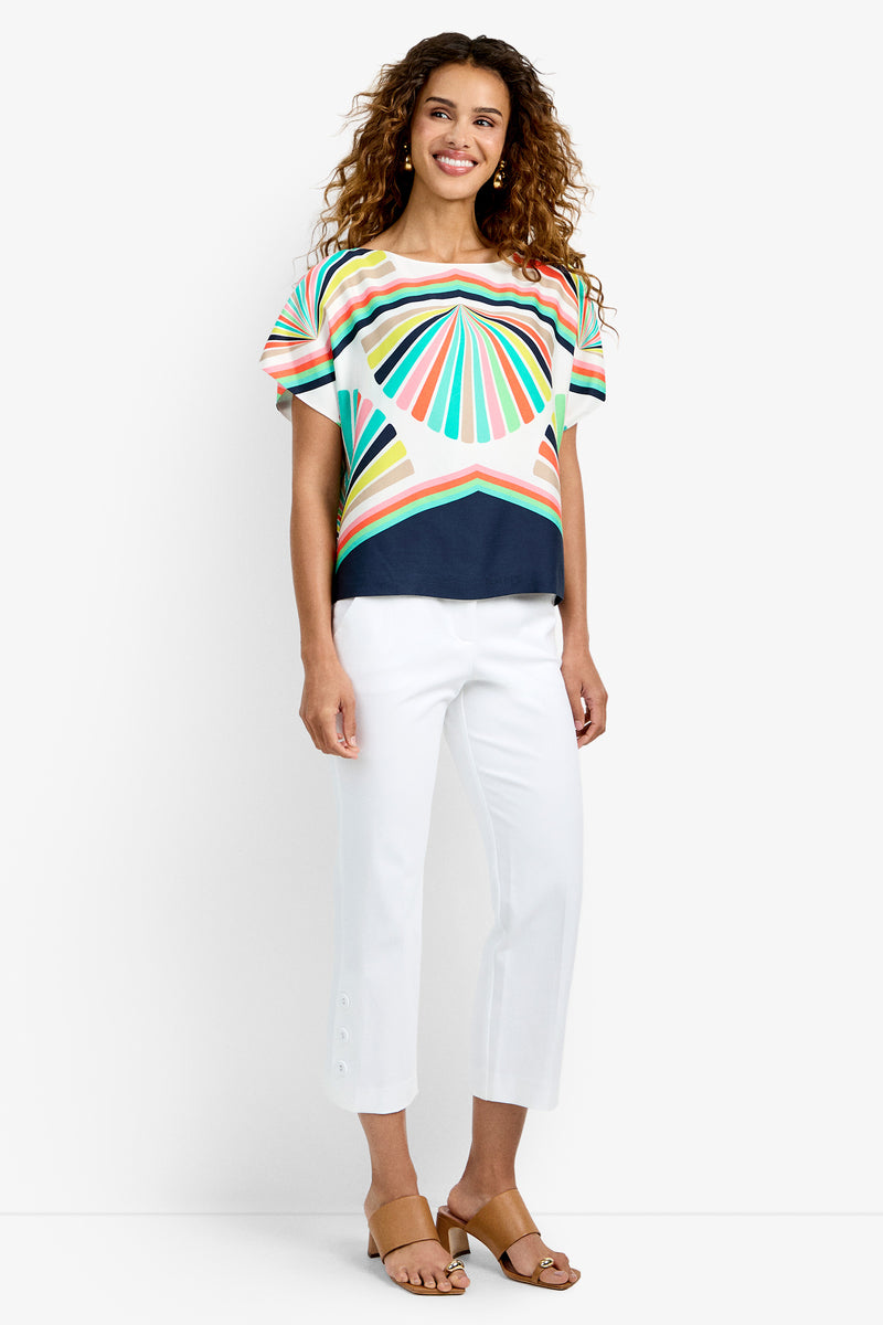 A woman stands smiling in the Breezy Top, a colorful geometric-patterned short-sleeve shirt, paired with white cropped pants with hem buttons and tan sandals, against a plain white background.
