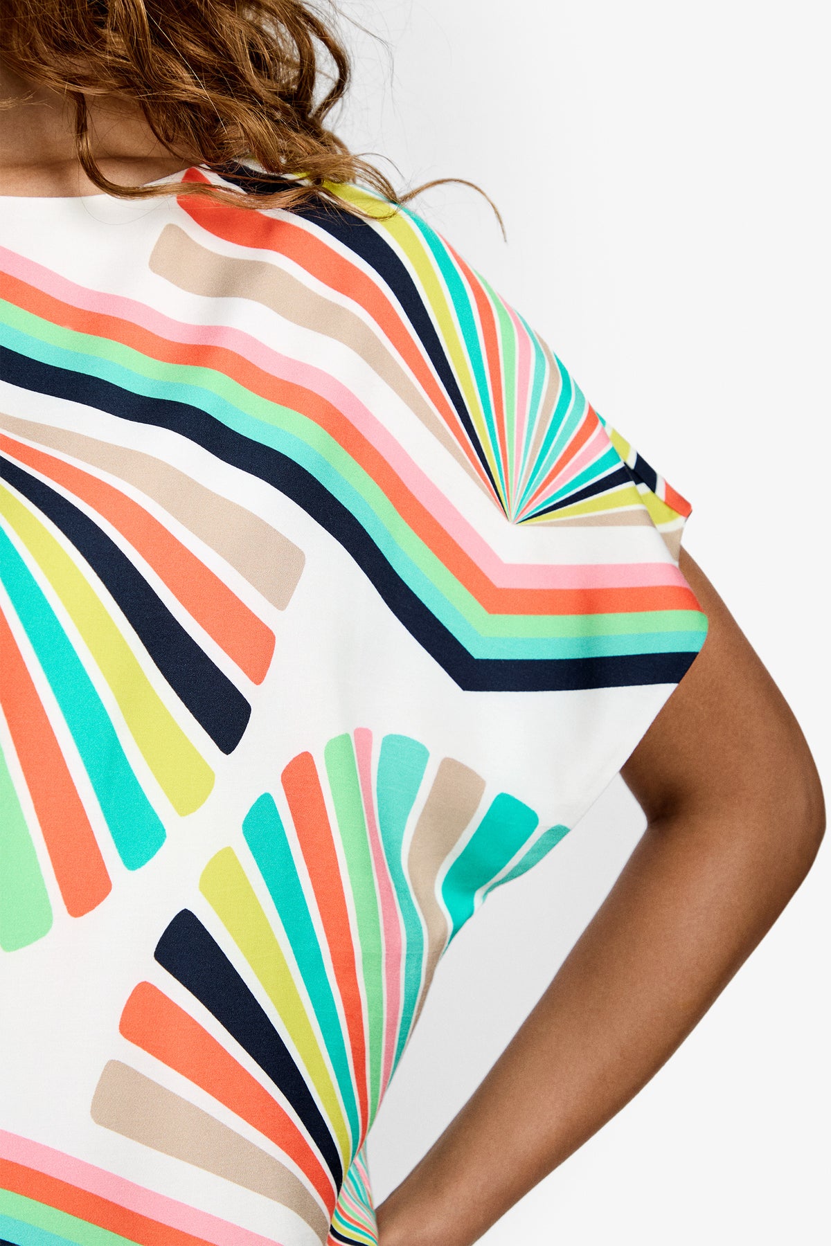 Close-up of a person wearing the Breezy Top, featuring colorful geometric fan-shaped patterns in orange, teal, navy, lime green, and beige. The white top is partially shown with a bent arm and some hair visible.