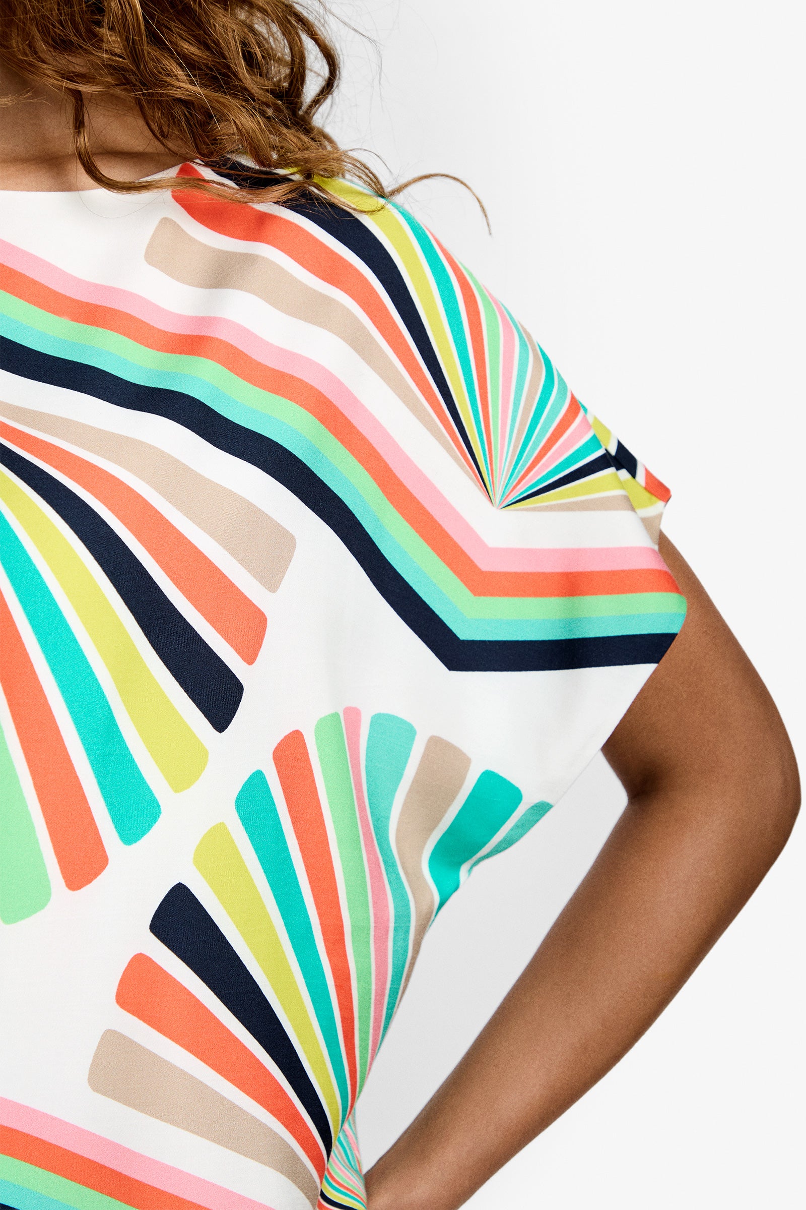 Close-up of a person wearing the Breezy Top, featuring colorful geometric fan-shaped patterns in orange, teal, navy, lime green, and beige. The white top is partially shown with a bent arm and some hair visible.