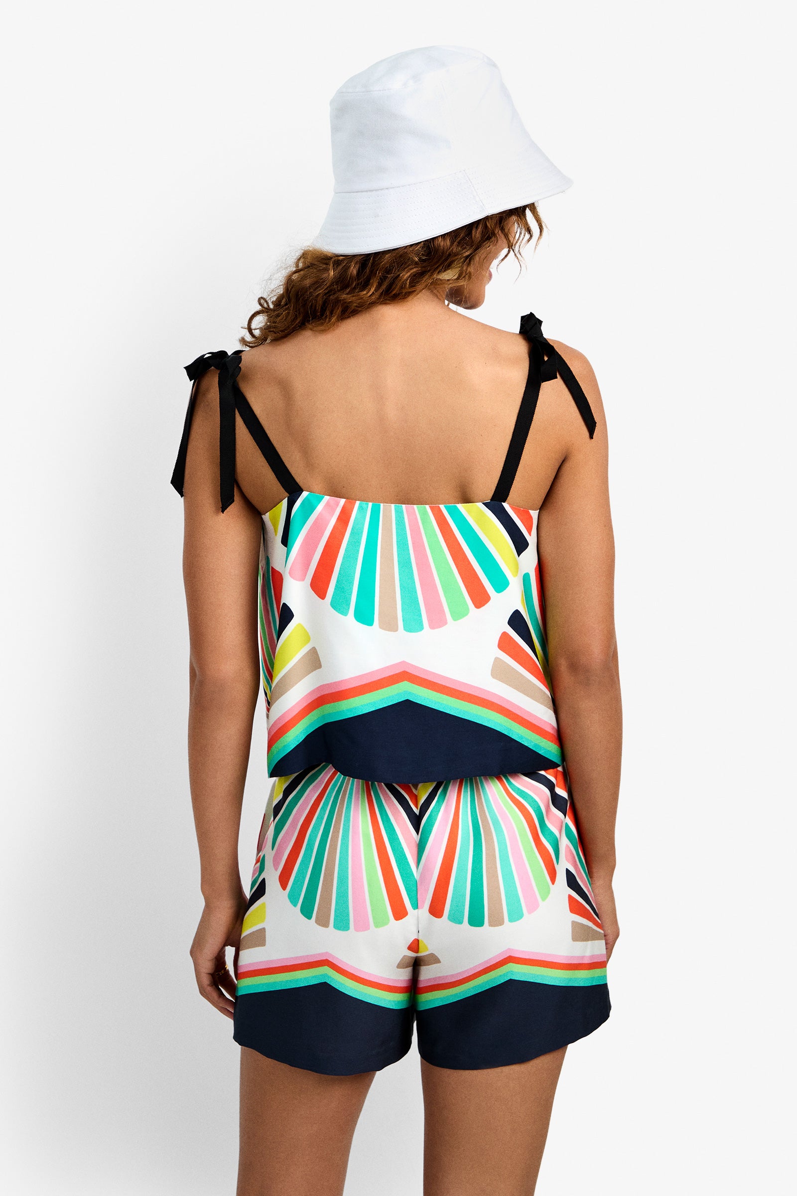 A woman faces away wearing a white bucket hat and the Marella Top—a colorful, geometric-patterned tank with black shoulder ties, multicolored stripes, and navy blue accents—paired with matching shorts.