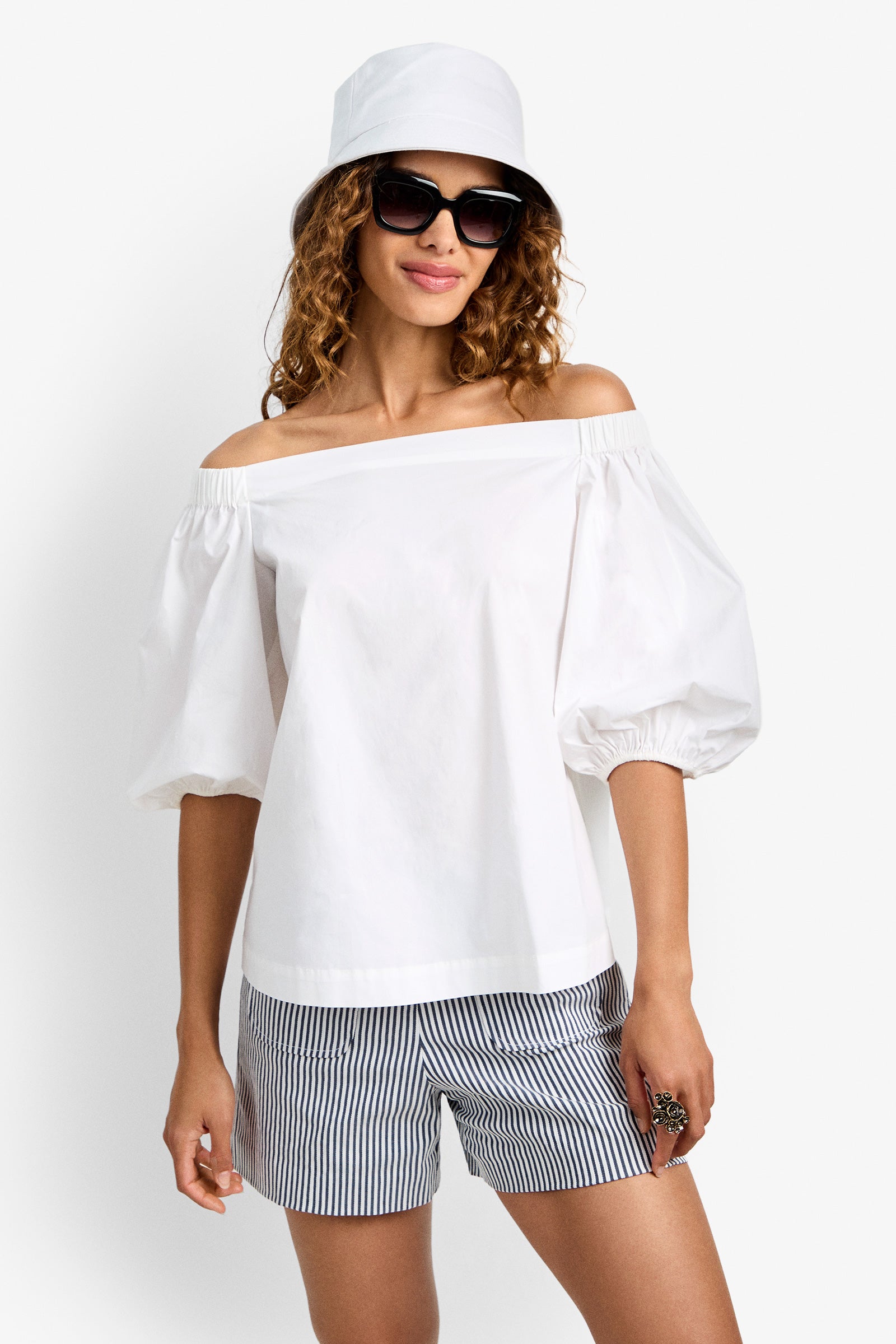 A woman smiles against a white background, wearing the Cove Top—a white off-the-shoulder design with puff sleeves—paired with black-and-white striped shorts, a white bucket hat, and large black sunglasses.