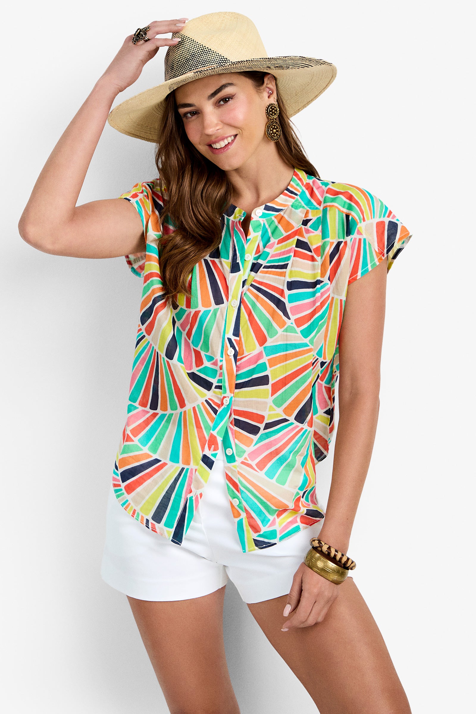 A woman smiles against a plain light background, wearing the Krill Top—a colorful geometric short-sleeve button-up blouse—paired with a wide-brimmed straw hat, white shorts, gold earrings, bracelets, and a ring.