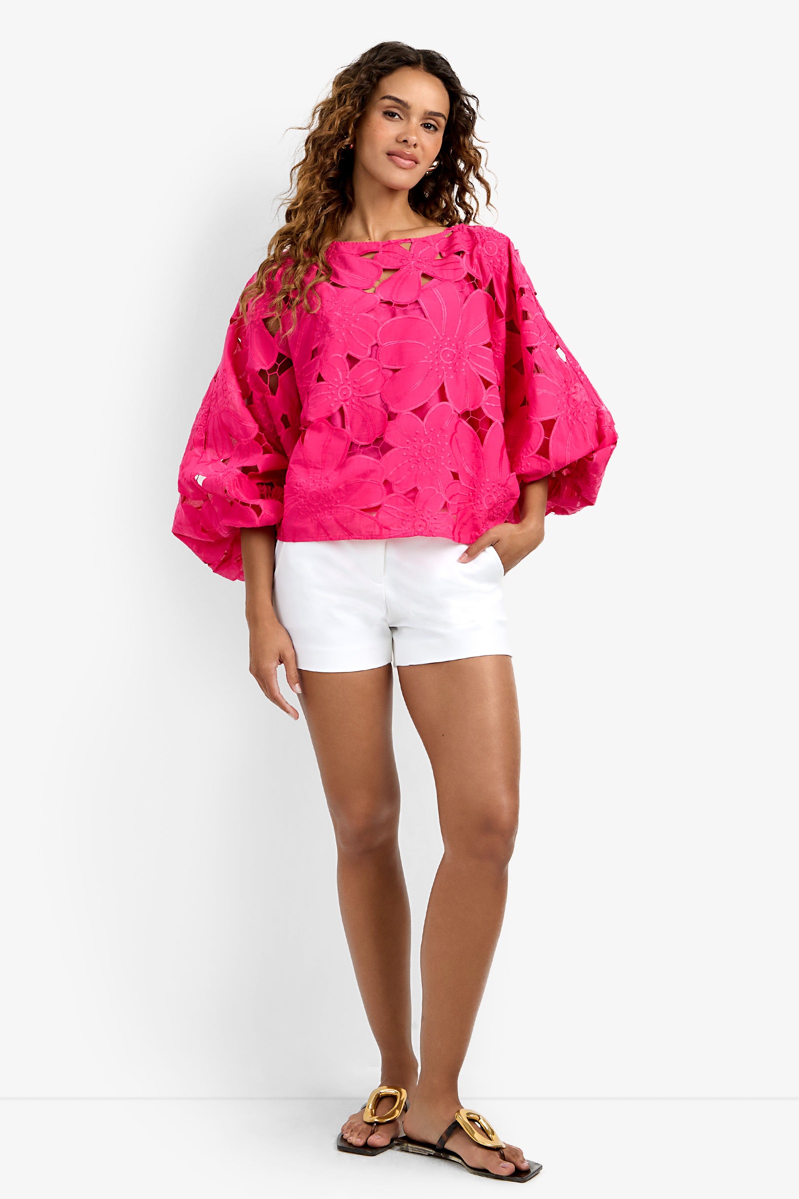 A woman with long curly hair poses in the Coastal Top, a bright pink floral-patterned oversized blouse with wide sleeves, paired with white shorts and black sandals against a plain white background.