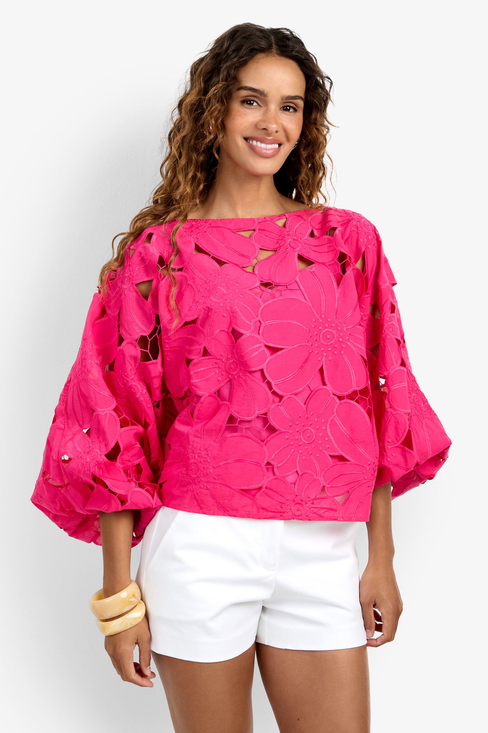 A woman with wavy hair and chunky bangles smiles while wearing the Coastal Top—a bright pink, floral-patterned, loose blouse with wide sleeves—paired with white shorts, posing against a plain background.