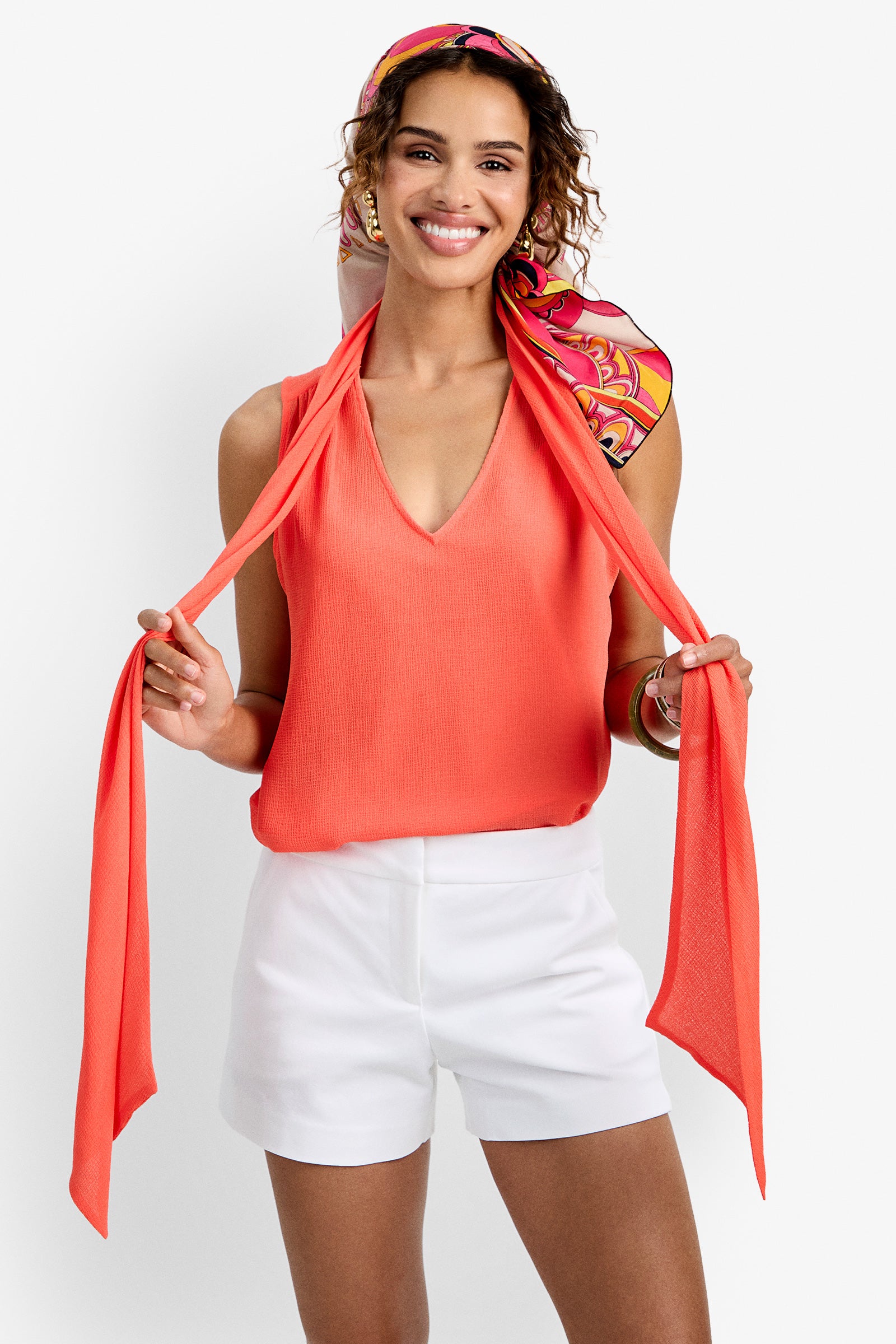 A woman with curly hair smiles while holding a colorful scarf on her head, wearing the sleeveless coral Alora Top and white shorts. She accessorizes with hoop earrings and bracelets against a white background.