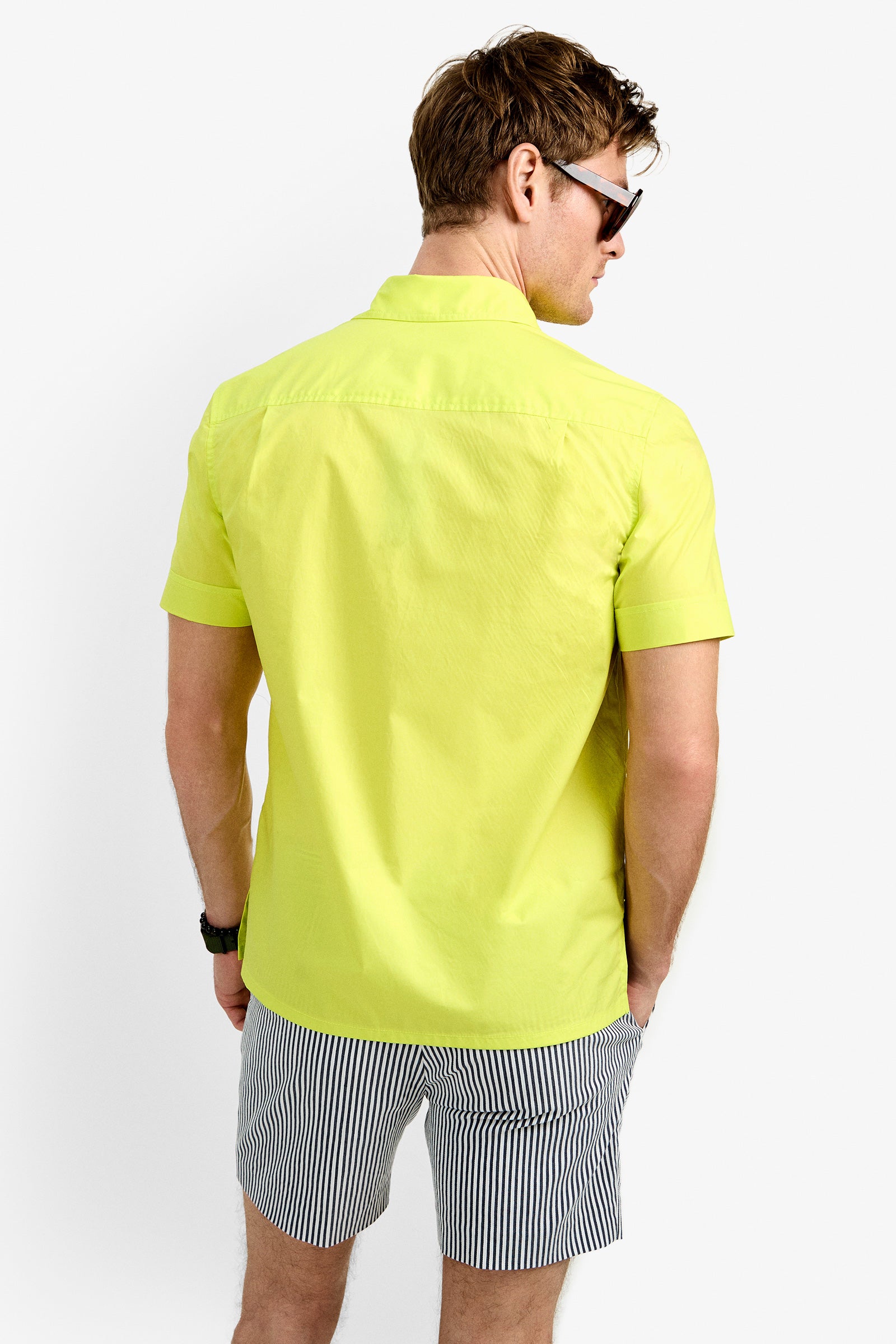 A man wearing the Will Shirt in bright yellow stands facing away, looking over his shoulder, styled with sunglasses, a watch, and black-and-white striped shorts against a plain white background.