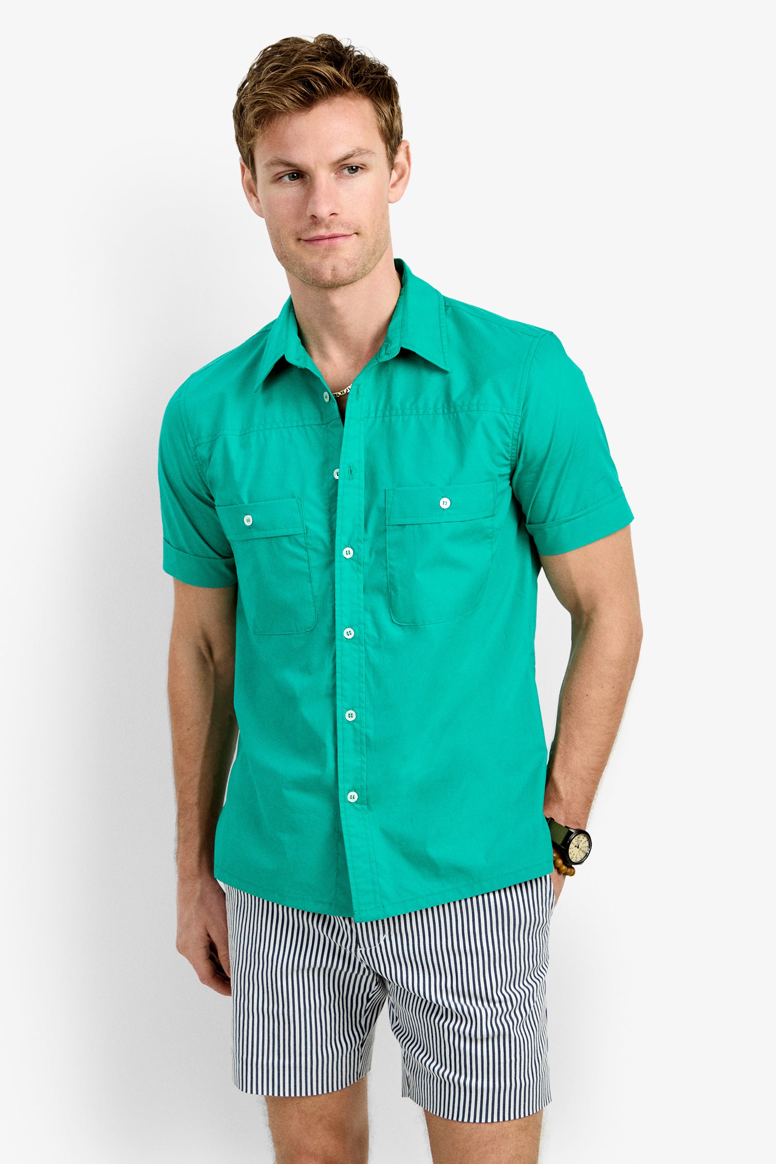 A man models the Will Shirt, a bright turquoise short-sleeve button-up, paired with blue-and-white striped shorts against a plain white background, looking slightly to the side.