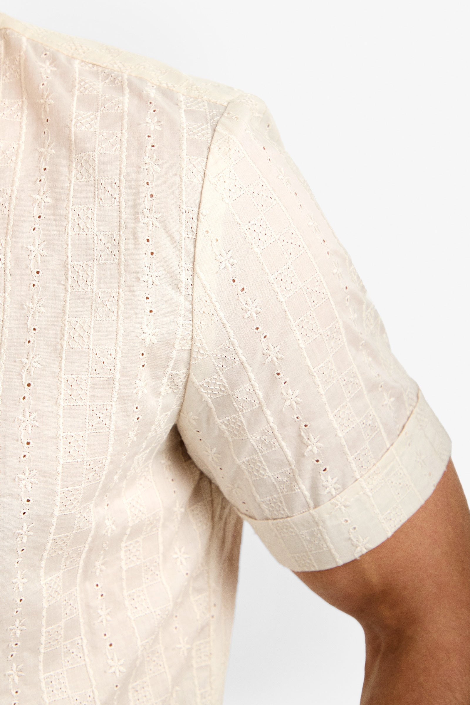 Close-up of a person wearing the Branson Shirt, a light beige short-sleeve top featuring embroidered geometric and floral patterns. The textured, slightly sheer fabric highlights detailed stitching on the sleeve and shoulder.