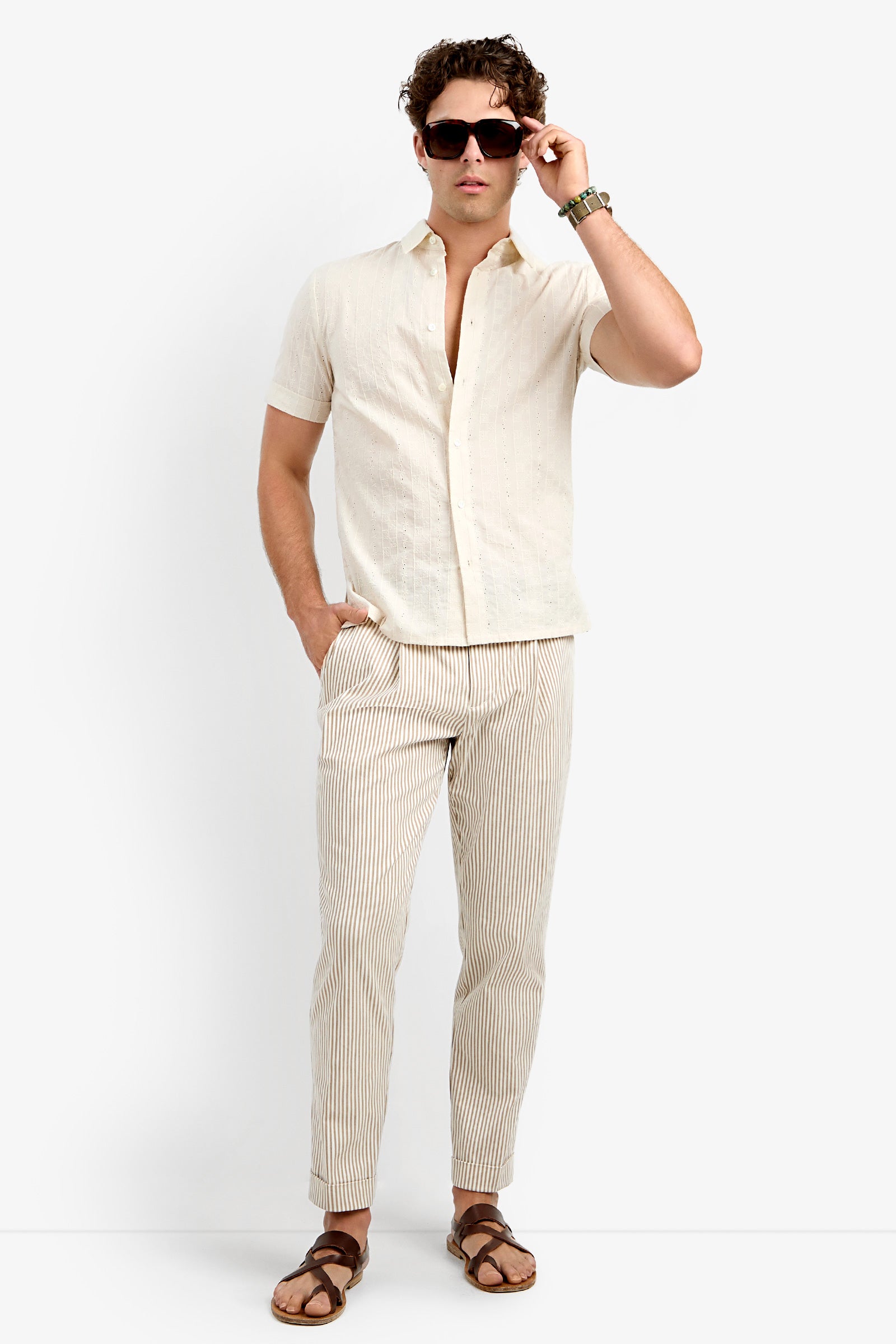 A man wears the Branson Shirt, a short-sleeve beige button-up, paired with tan and white striped pants and brown sandals as he poses with one hand in his pocket against a plain white background.