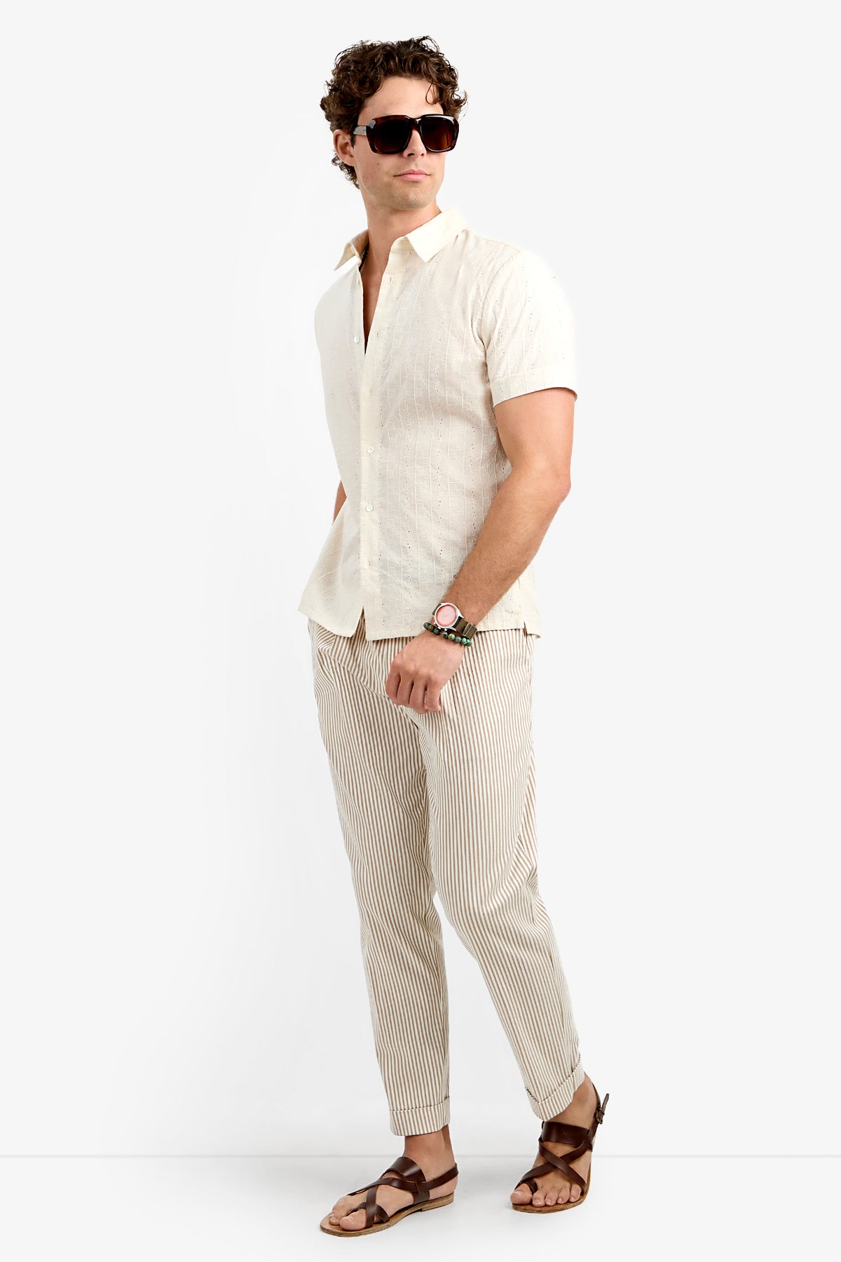A man wearing sunglasses, the Branson Shirt (a short-sleeve off-white button-up), light pinstriped pants, and brown sandals stands against a plain white background.