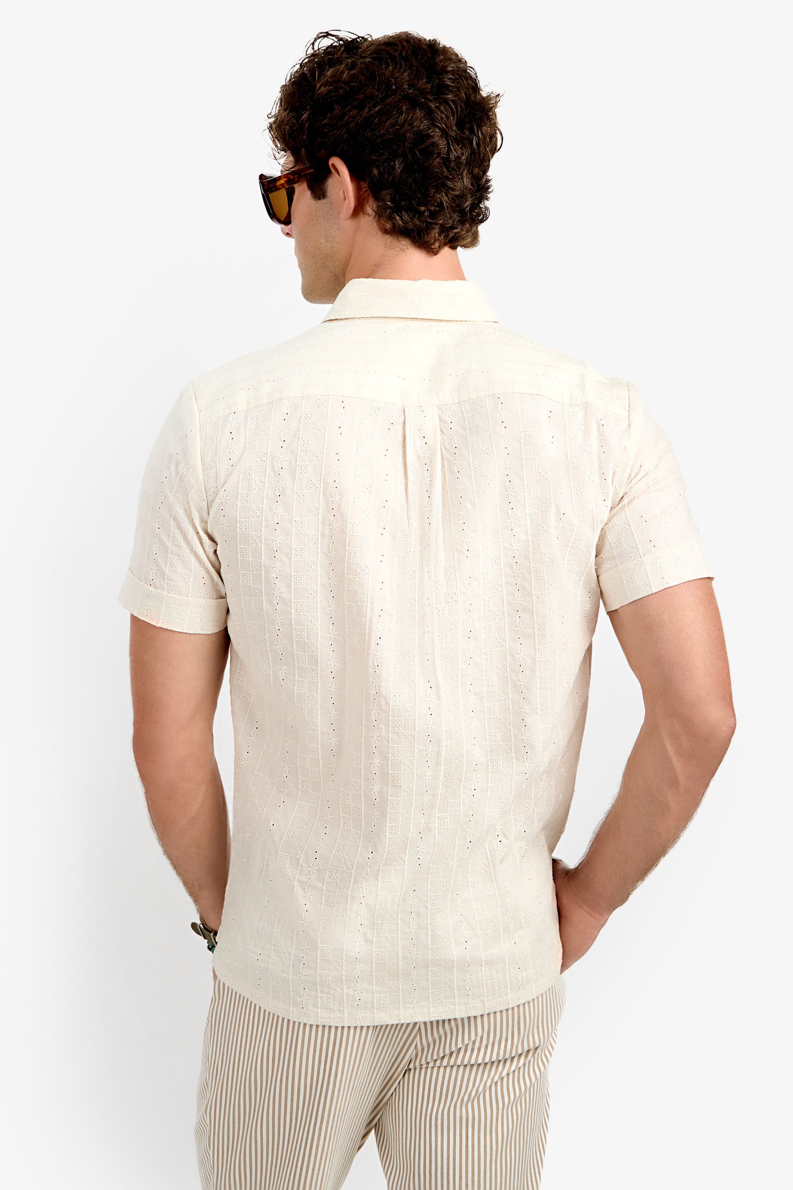 A man with curly hair and sunglasses is seen from behind wearing the Branson Shirt, a light beige short-sleeve shirt with a subtle pattern, paired with beige striped pants against a plain white background.