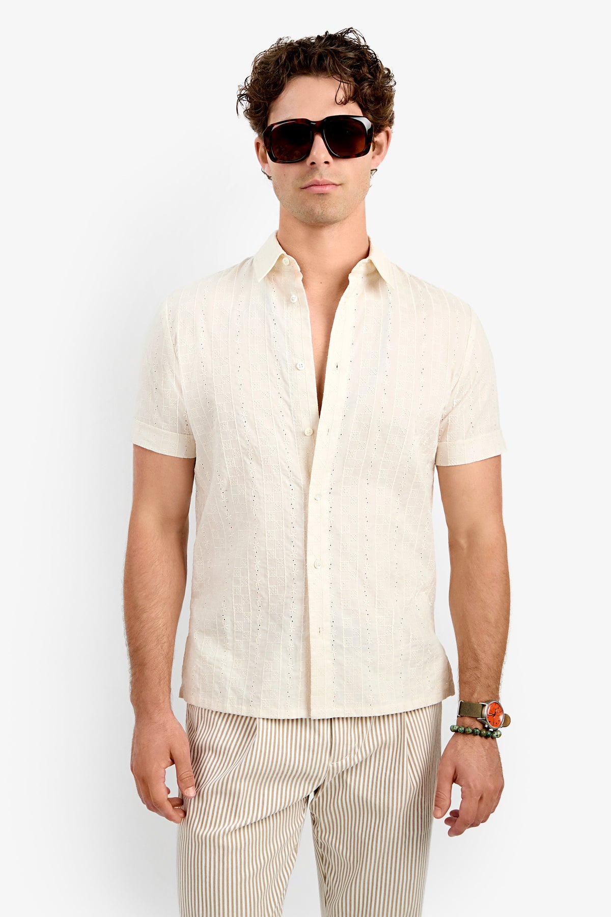 A person with curly hair stands facing forward, wearing large dark sunglasses, bracelets on their left wrist, the short-sleeve patterned Branson Shirt in cream, and light beige striped pants.