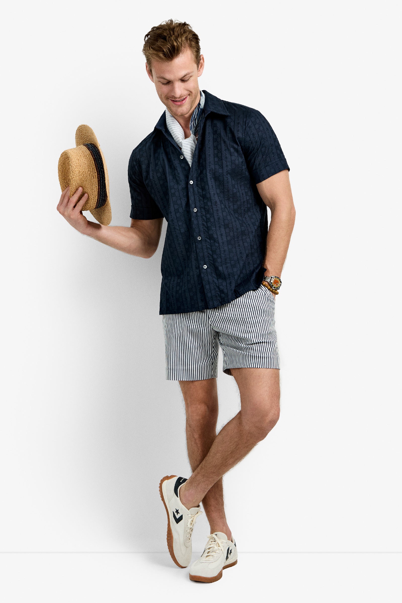 A man smiles against a white background, wearing the Branson Shirt in INK over a white shirt, striped shorts, white sneakers, and holding a straw hat in his left hand.