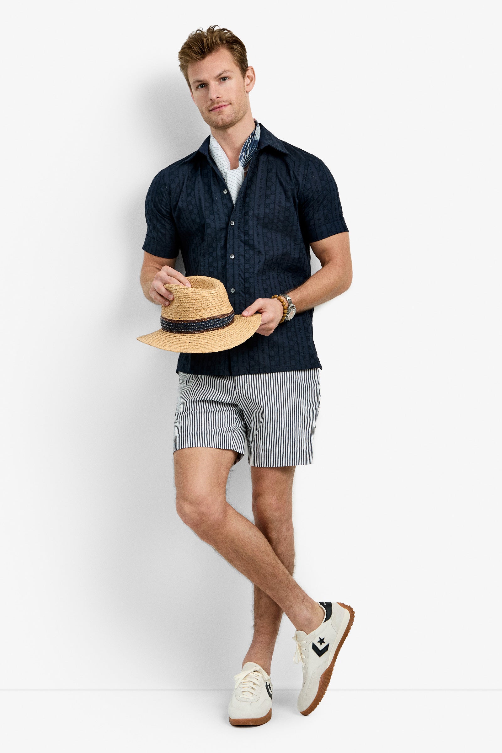 A man with light brown hair stands in a relaxed pose against a white background, wearing the Branson Shirt in navy over a light blue shirt, striped shorts, white sneakers, and holding a straw hat.
