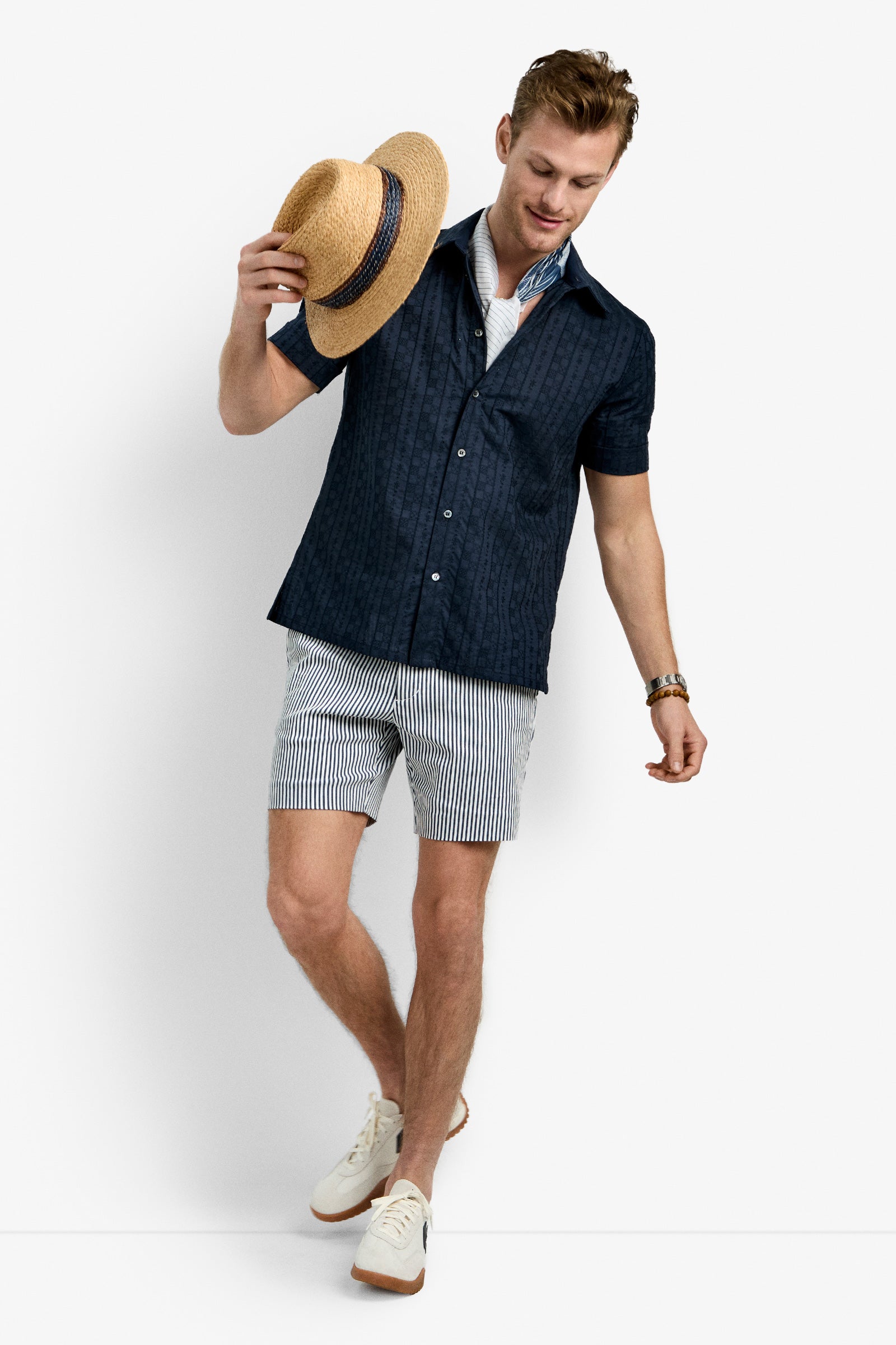 A man in the Branson Shirt, striped shorts, white sneakers, and a bracelet holds a straw hat and smiles while looking down against a plain white background.