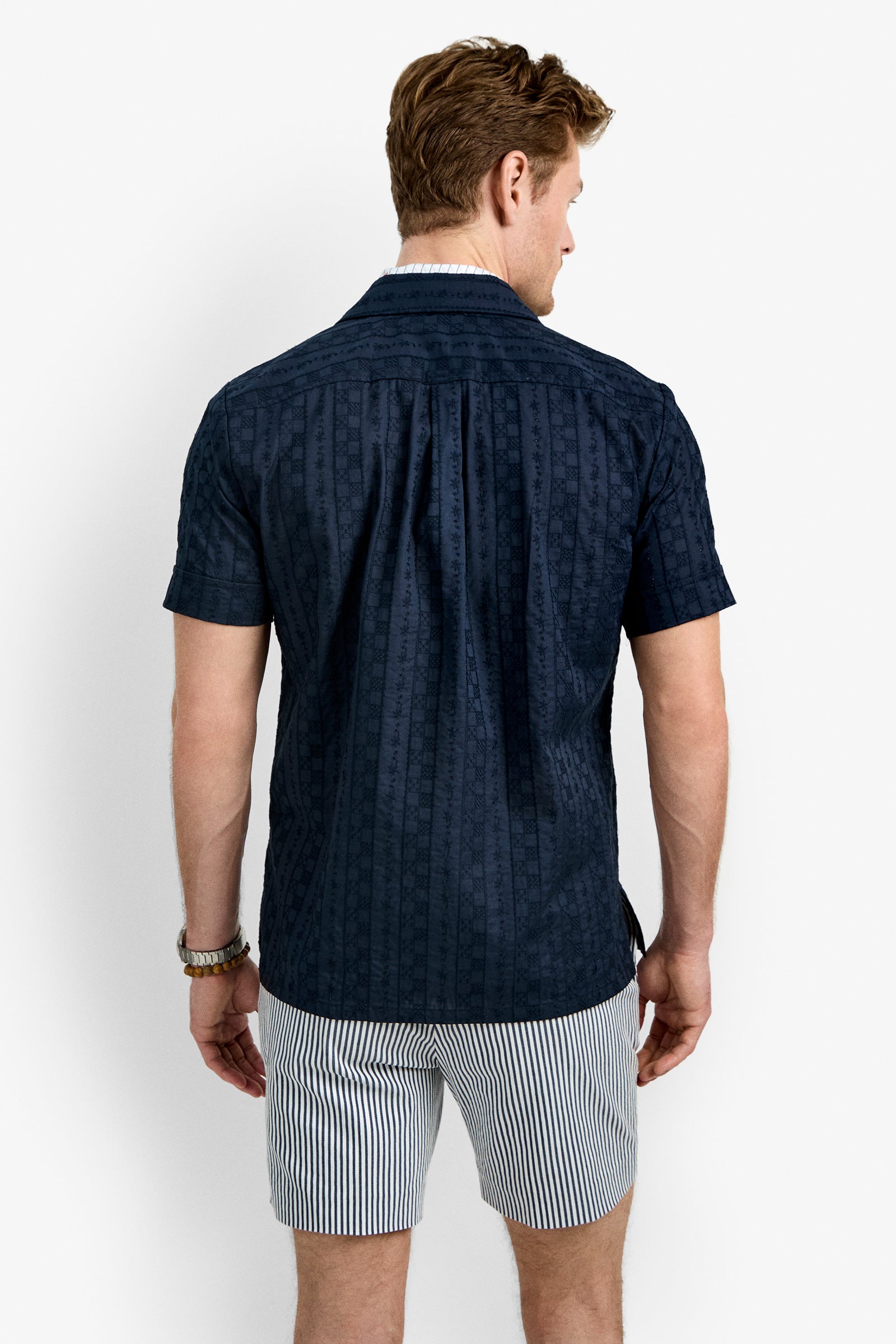 A man with light brown hair and a watch on his left wrist faces away from the camera, wearing the Branson Shirt— a navy short-sleeve button-up — paired with white shorts featuring vertical blue stripes.