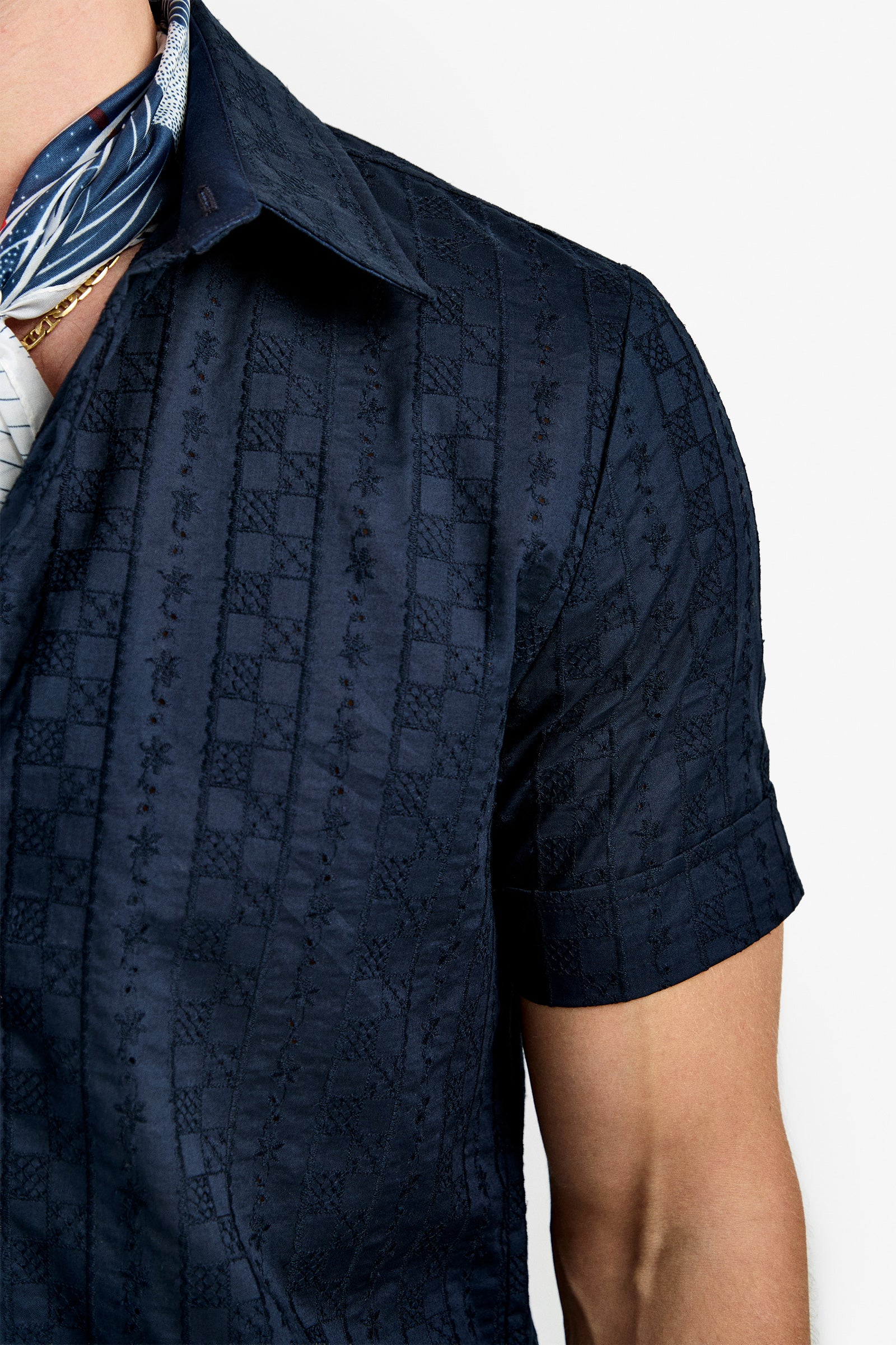 A close-up of a person wearing the Branson Shirt—a dark blue, short-sleeve, textured button-up with a subtle pattern—paired with a blue and white patterned scarf and gold chain necklace. Background is plain white.