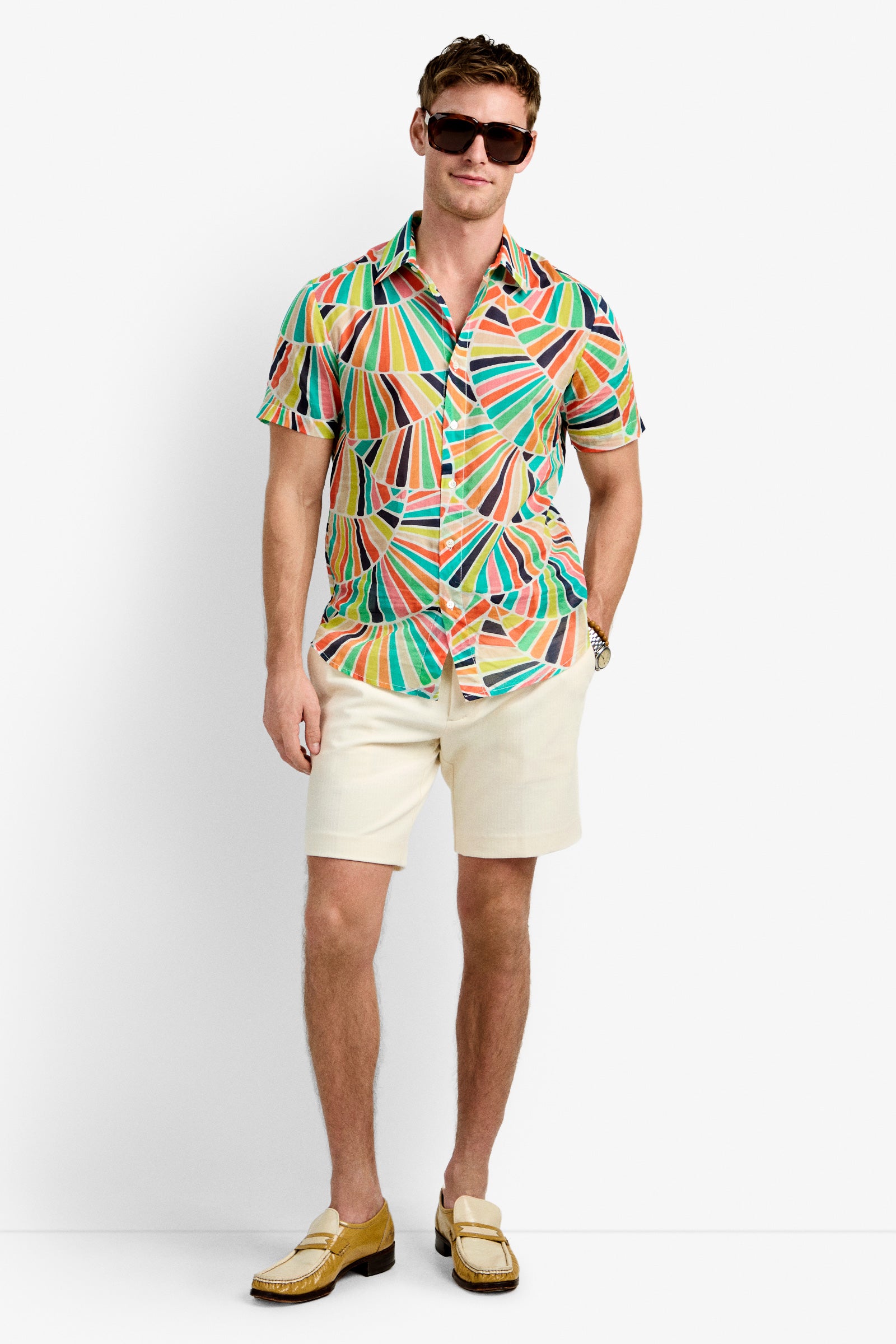 A man wearing dark sunglasses, the Slim Jim Shirt, cream shorts, and tan loafers stands against a plain white background.